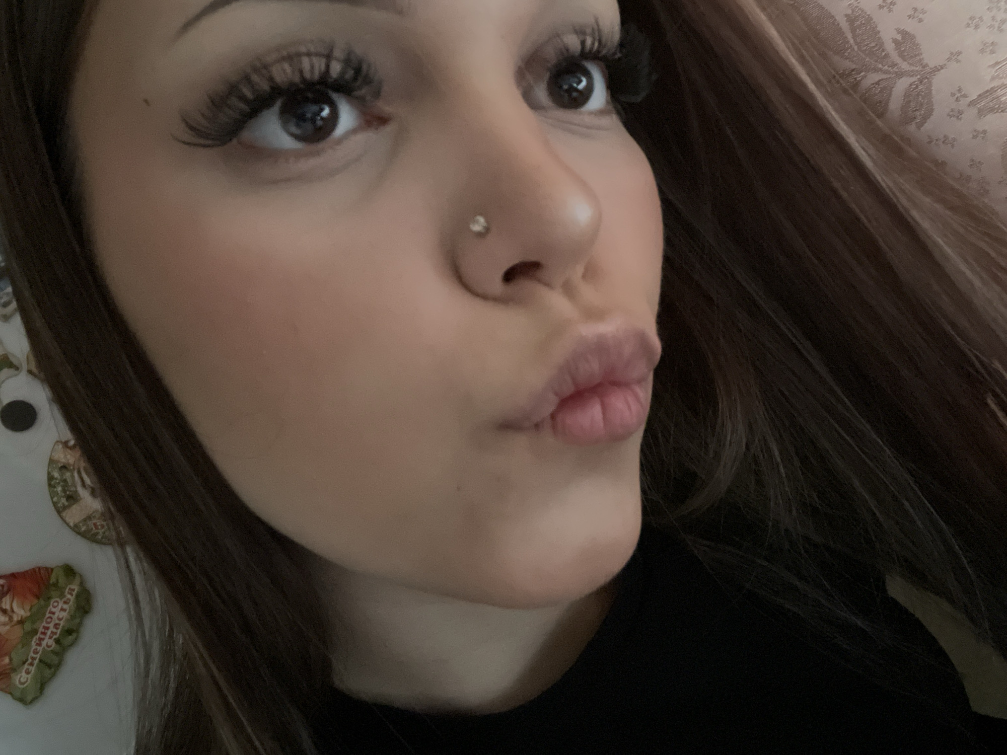 A close-up portrait captures a young woman from the chest up, angled slightly to her right, with her gaze directed upwards. She has long, dark brown hair, dark eyes, and prominent eyelashes. A small, clear stud is visible in her left nostril. Her lips are pursed in a "kissing" or "pouting" expression, conveying a playful or affectionate mood. She is wearing a dark, possibly black, top.
The background on the right side is a light-colored, patterned fabric or wallpaper. On the far left, a light-colored surface, possibly a refrigerator, is visible with a few decorations. One of these decorations is a heart-shaped magnet with Cyrillic text. The visible text on the magnet reads "СЕМЕЙНОГО СЧАСТЬЯ", which translates to "FAMILY HAPPINESS". The lighting appears to be indoor and soft. The overall scene suggests a casual, personal photo, likely taken indoors in a home setting. Given the location context, these cues are consistent with a setting in Russia.