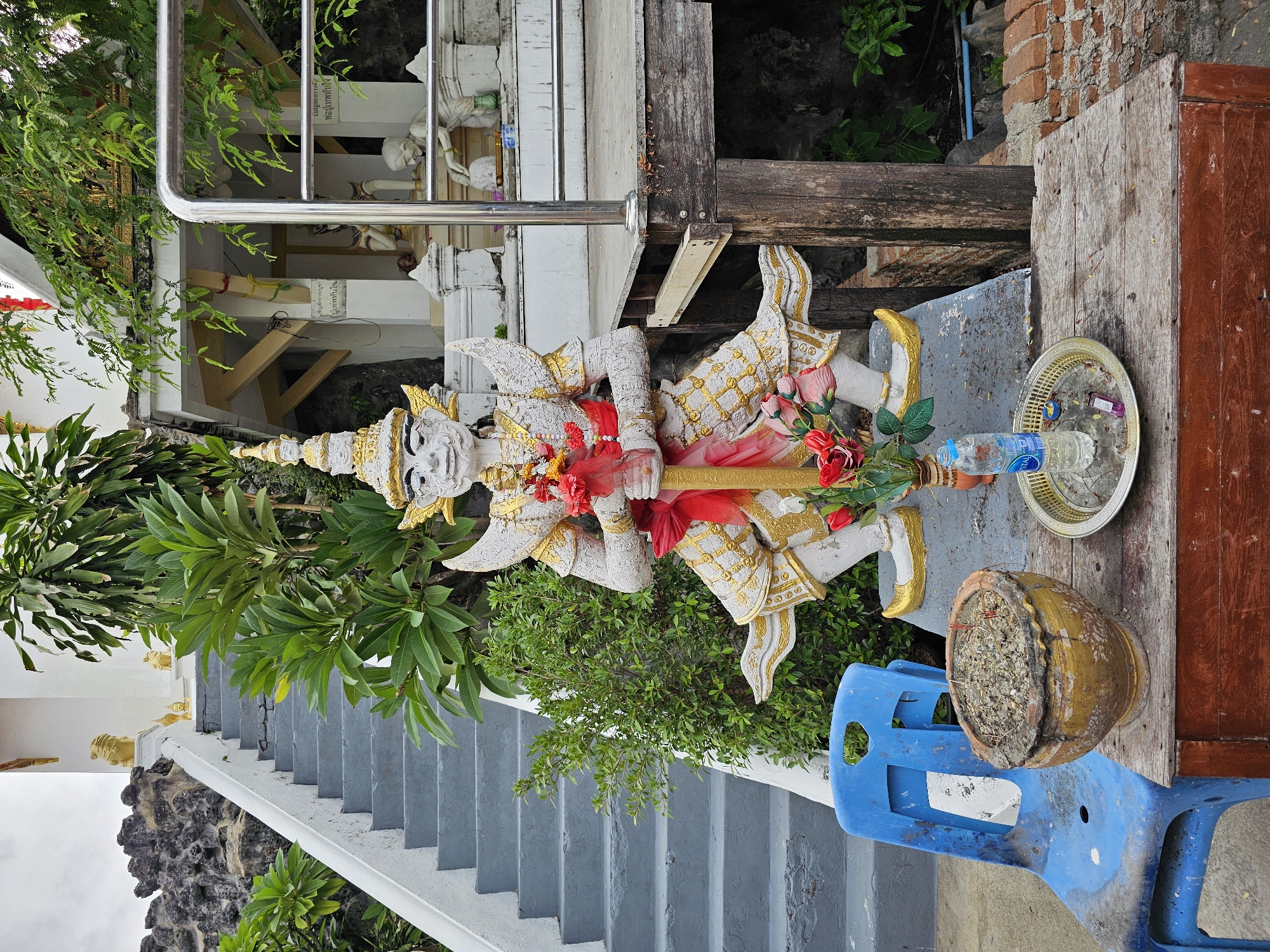 This image captures a scene in Bangkok, Thailand, likely at a temple or shrine. The central focus is a large, ornate statue of a mythical guardian figure, characterized by its stern expression, elaborate headdress, and decorative white and gold attire. The statue holds a staff or sword-like object in its hands. In front of the statue, on a weathered wooden table, are several offerings: a small vase of artificial red flowers, a metal tray with a bottle of water and a cigarette lighter, and a clay pot filled with ashes and incense sticks. To the left of the statue, a weathered blue plastic chair sits beside a large, decorative clay pot. Behind the statue, a grey concrete staircase ascends, flanked by lush green foliage. Further in the background, a small shrine structure with white walls and gold trim is visible, supported by wooden beams. The overall environment suggests an outdoor religious or cultural site, possibly on a slightly overcast day given the lighting.