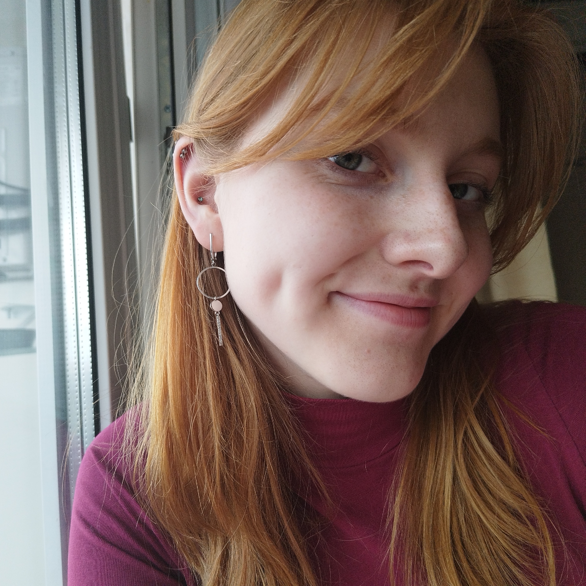 A close-up selfie shows a fair-skinned woman with freckles and reddish-blonde hair. She has dimples and is smiling. She is wearing a maroon turtleneck shirt. In her left ear, she has a cartilage piercing and a dangling silver earring with a circle and a smaller charm. She appears to be inside, next to a window. The view through the window shows a gray, wintry cityscape. Based on the style of the window frame, the buildings visible, and the wintry atmosphere, the location may be in a city like Yekaterinburg, Russia, however, without additional context such as identifying landmarks, this cannot be confirmed.
