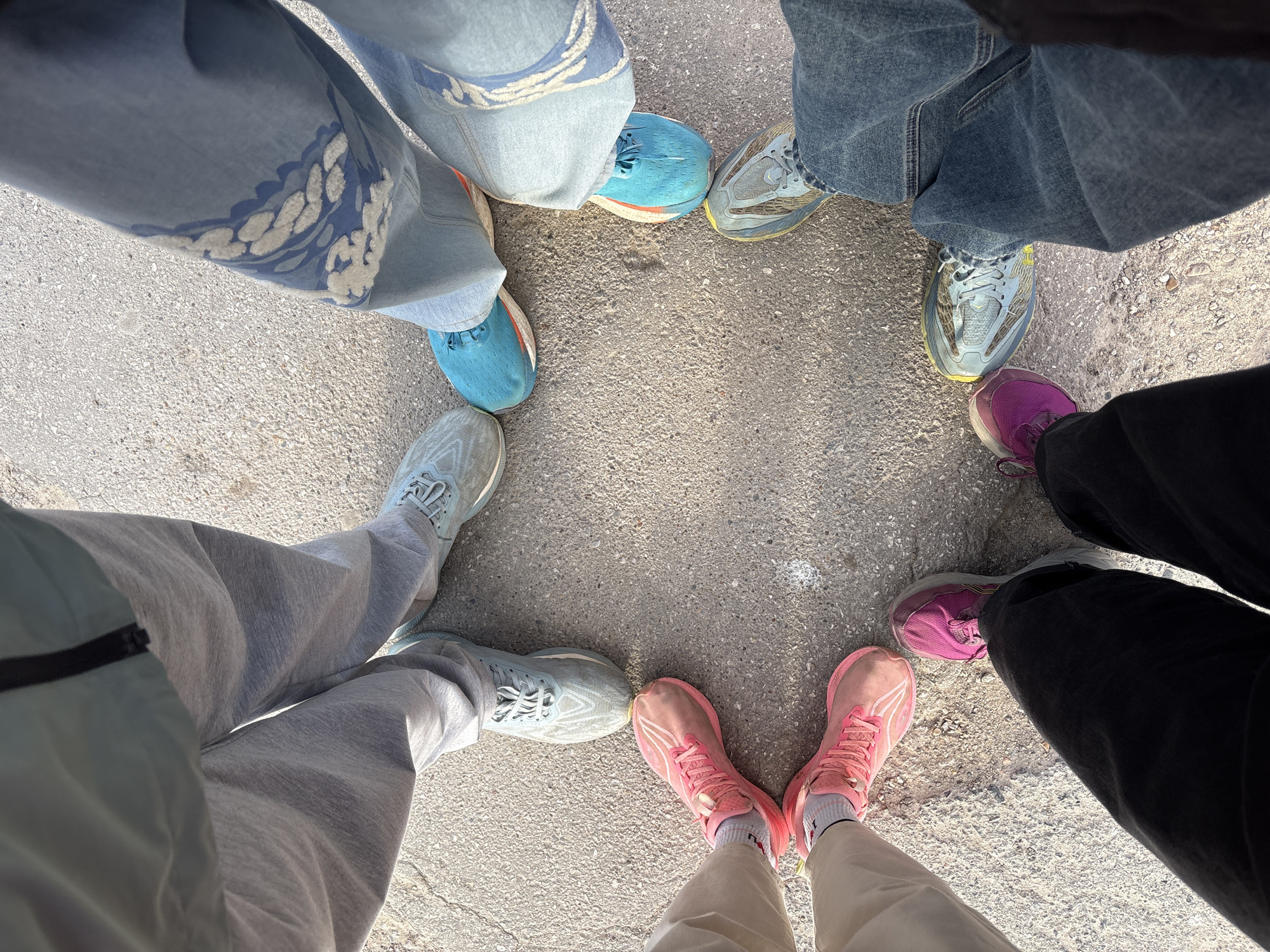 The image shows a circle of six people's feet and shoes, photographed from above. The shoes are various colors: blue, gray, pink, purple, and variations of light blue and gray. The people are wearing different colored pants: light blue jeans with a pattern, dark blue jeans, gray sweatpants, tan pants, and black pants. The setting appears to be a paved outdoor surface. The city of Perm, Russia, is not explicitly identifiable from the visual information provided.