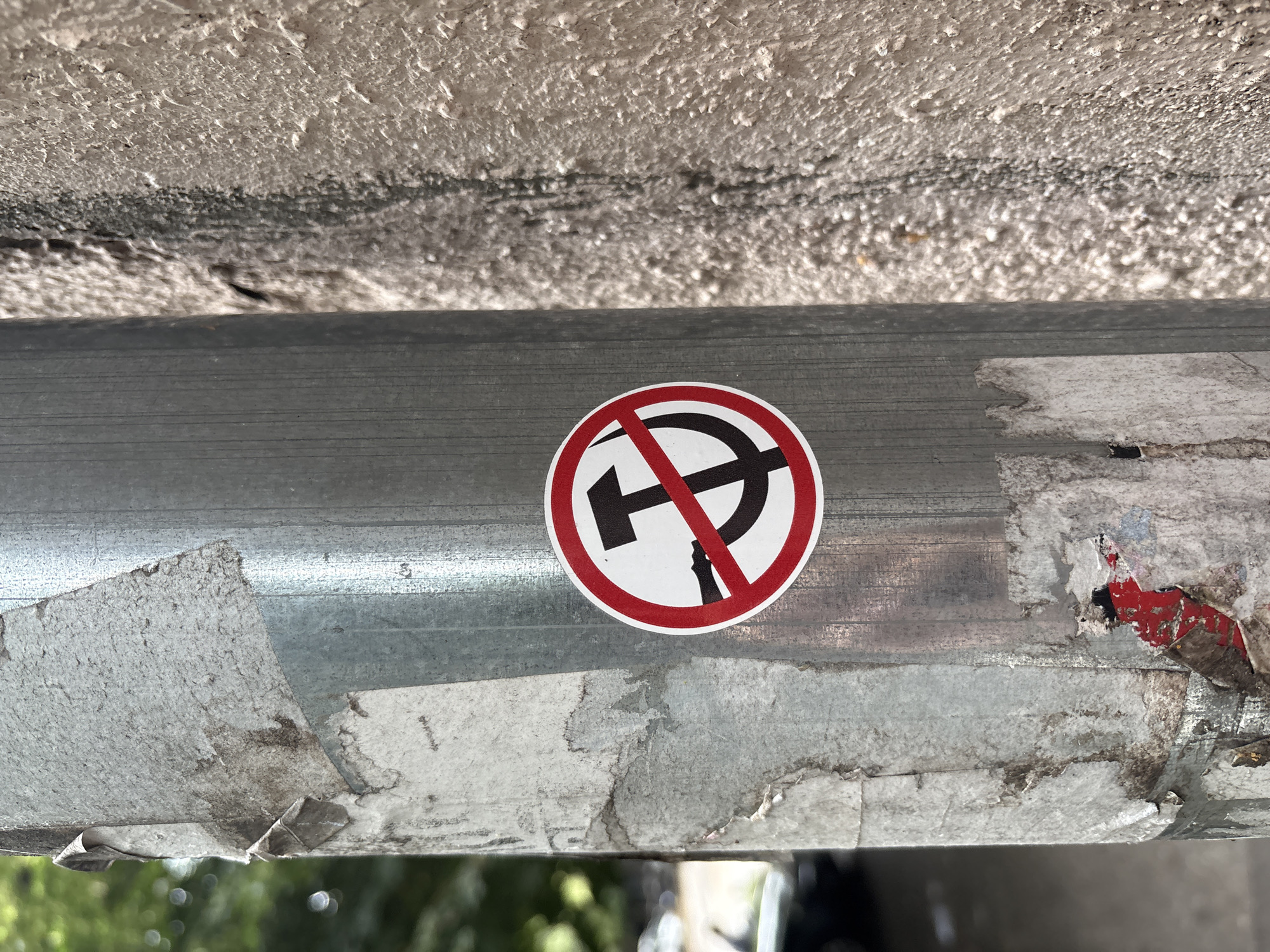 A close-up, eye-level, outdoor shot focuses on a silver metal pole, textured with peeling stickers and grime. Affixed to the pole is a circular sticker with a red border. Inside the border, a black hammer and sickle symbol is crossed out by a diagonal red line. To the right of the pole, a rough, beige, stucco wall creates a stark contrast. In the blurred background, green foliage and a dark car suggest an urban setting. This image was captured on a street in Belgrade, Serbia.