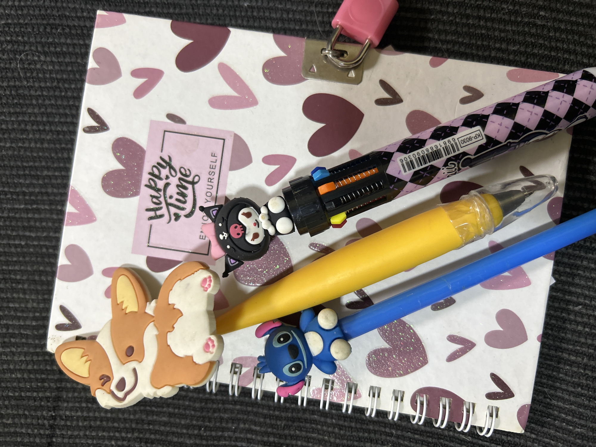 The image shows a close-up of a spiral-bound notebook with a white cover decorated with various shades of pink hearts. On the notebook, there are several objects including a yellow pen with a corgi charm, a blue pen with a Stitch charm, and a multi-colored pen with a Kuromi charm. A small pink padlock is attached to the right side of the notebook. The notebook and pens are placed on a dark, textured surface, likely a table or carpet. The scene is indoors and there are no people visible. There is no information in the image that indicates the location in the city of Buenos Aires, Argentina.
