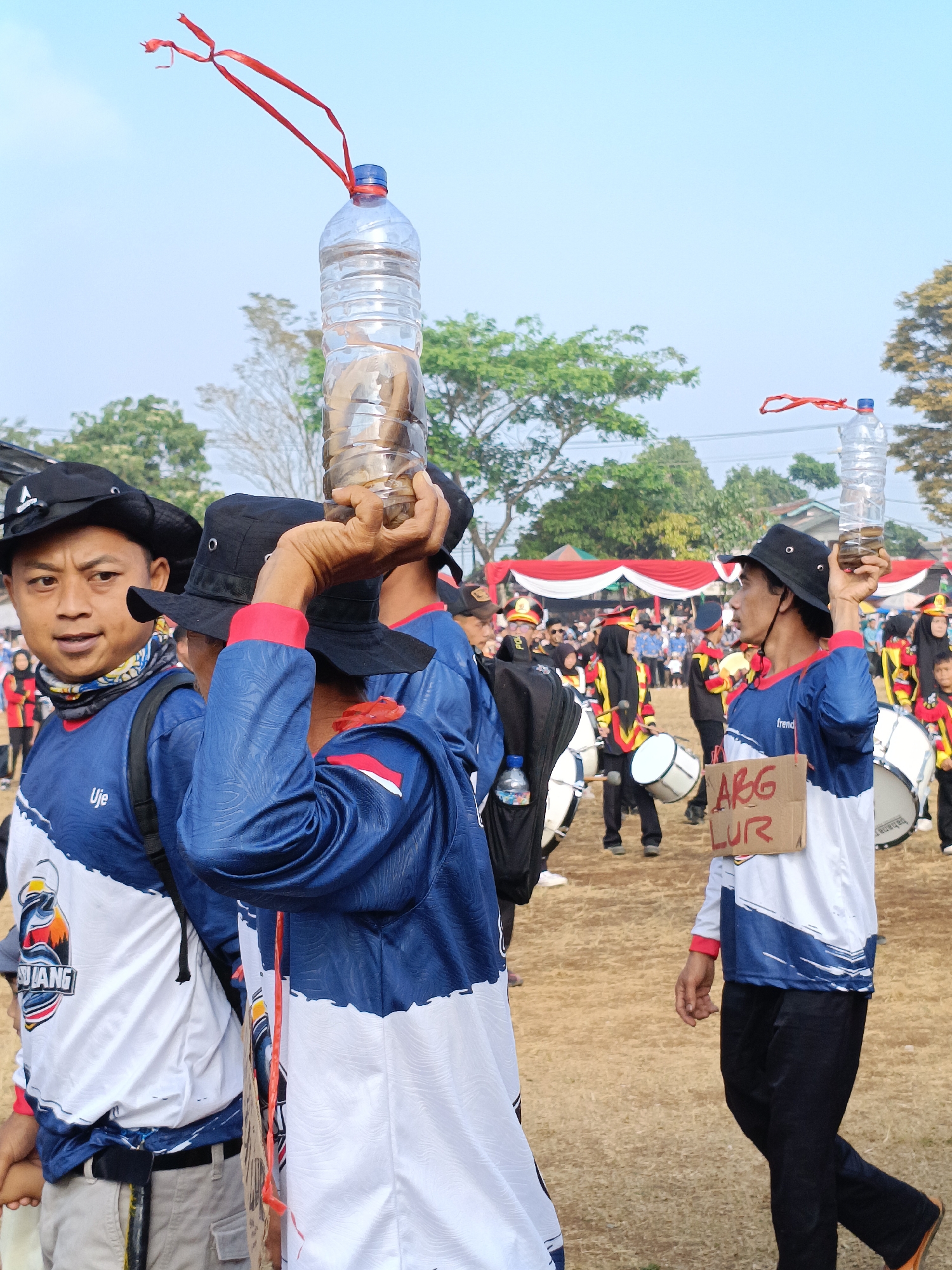 In Cibodas, Indonesia, a parade is underway on a dry, grassy field, marked by scattered trees and a clear blue sky. A group of individuals, some wearing black bucket hats and blue and white long-sleeved shirts with red accents, are participating. In the foreground, a man in a black hat holds a large plastic water bottle filled with a dark liquid and some objects, balanced on his head, with a red ribbon tied to its cap. Another individual in a similar outfit stands next to him, also wearing a black hat. Further back, other participants are visible, some carrying musical instruments like drums. A few are also holding a cardboard sign with red lettering that reads "ARG LUR". In the background, tents with red and white banners are set up, suggesting a public event.