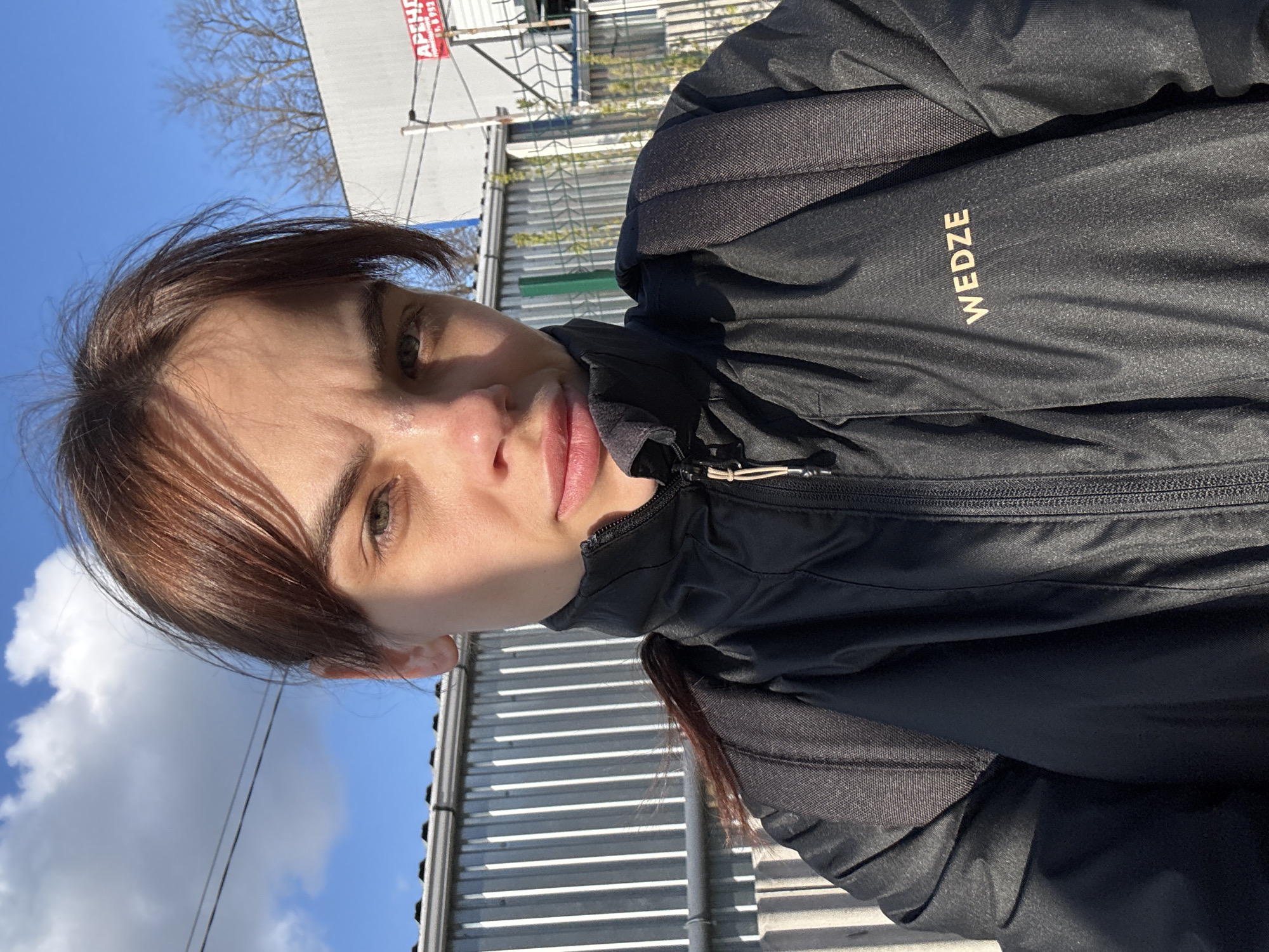 A woman is standing outside on a sunny day. She has light skin, brown hair with bangs, and is wearing a black Wedze jacket. Behind her, there is a blue sky with clouds, a building, and trees. The building appears to have a metal exterior, and there is a sign in Russian visible. The location is likely in Rostov-on-Don, Russia, as specified.