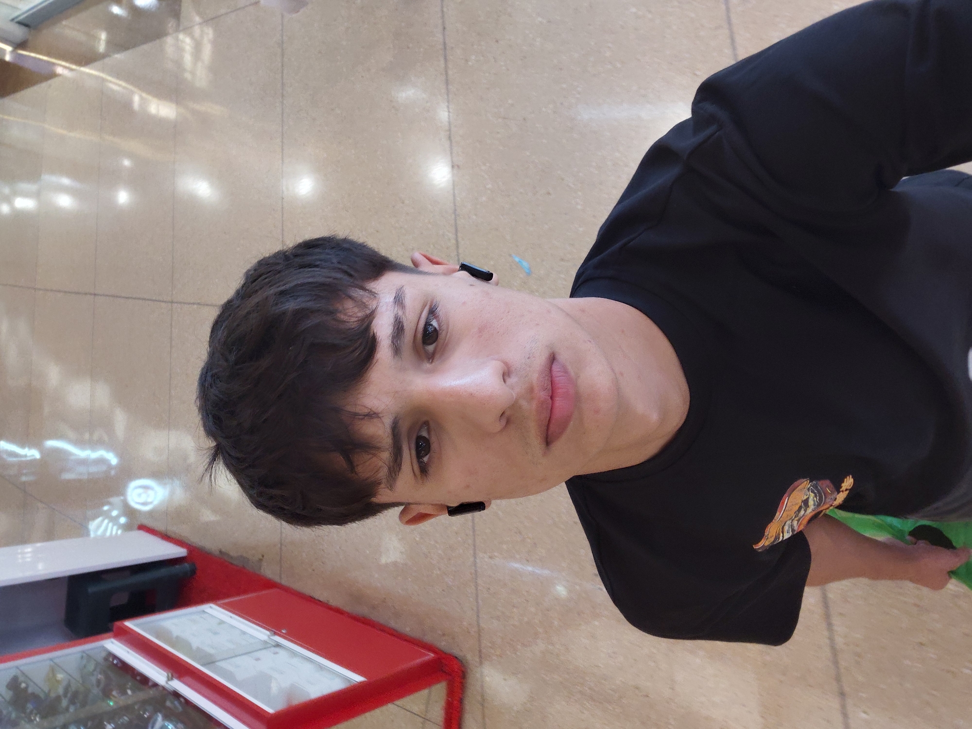 A young man with dark, tousled hair and wearing a black t-shirt with a colorful graphic on the left chest stands in what appears to be a well-lit indoor public space with a tiled floor. He is wearing black wireless earbuds in both ears. In the background to the left, a red and white display case is visible, possibly in a retail setting. The flooring is light-colored and reflective, with patterns of light suggesting overhead illumination. The overall impression is of a candid shot taken in a commercial or public walkway.