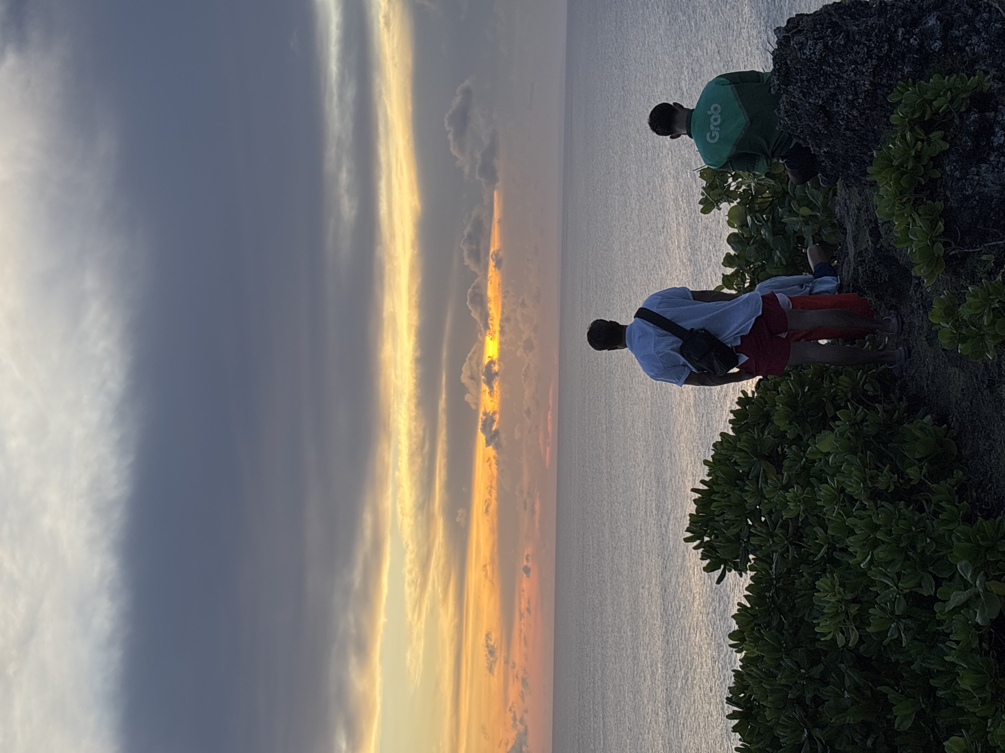 Two men are on a cliff overlooking the ocean at sunset. One man wears a green "Grab" shirt, and the other wears a white shirt and red shorts. They stand among green bushes and rocks. The sky transitions from dark clouds to a vibrant orange sunset. This is in Uluwatu, Indonesia.