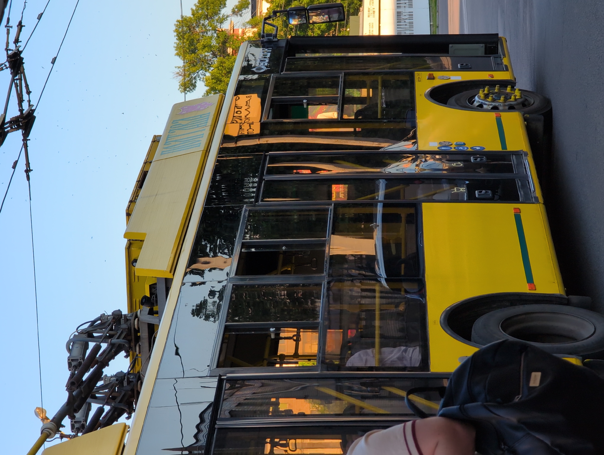 A side view of a yellow trolleybus is captured on a clear day in an urban environment. The trolleybus, with its dark tinted windows reflecting the surroundings, is equipped with a pantograph system on its roof, connected to overhead power lines. The front of the trolleybus is not fully visible, but the side displays the vehicle's number and various symbols. The backdrop includes a lush green tree and a building, suggesting a street setting. The ground is paved, indicating a road or a bus stop area. A person is partially visible on the left, holding a black backpack, preparing to board or disembark the vehicle. The overall impression is that of a functional public transport scene.