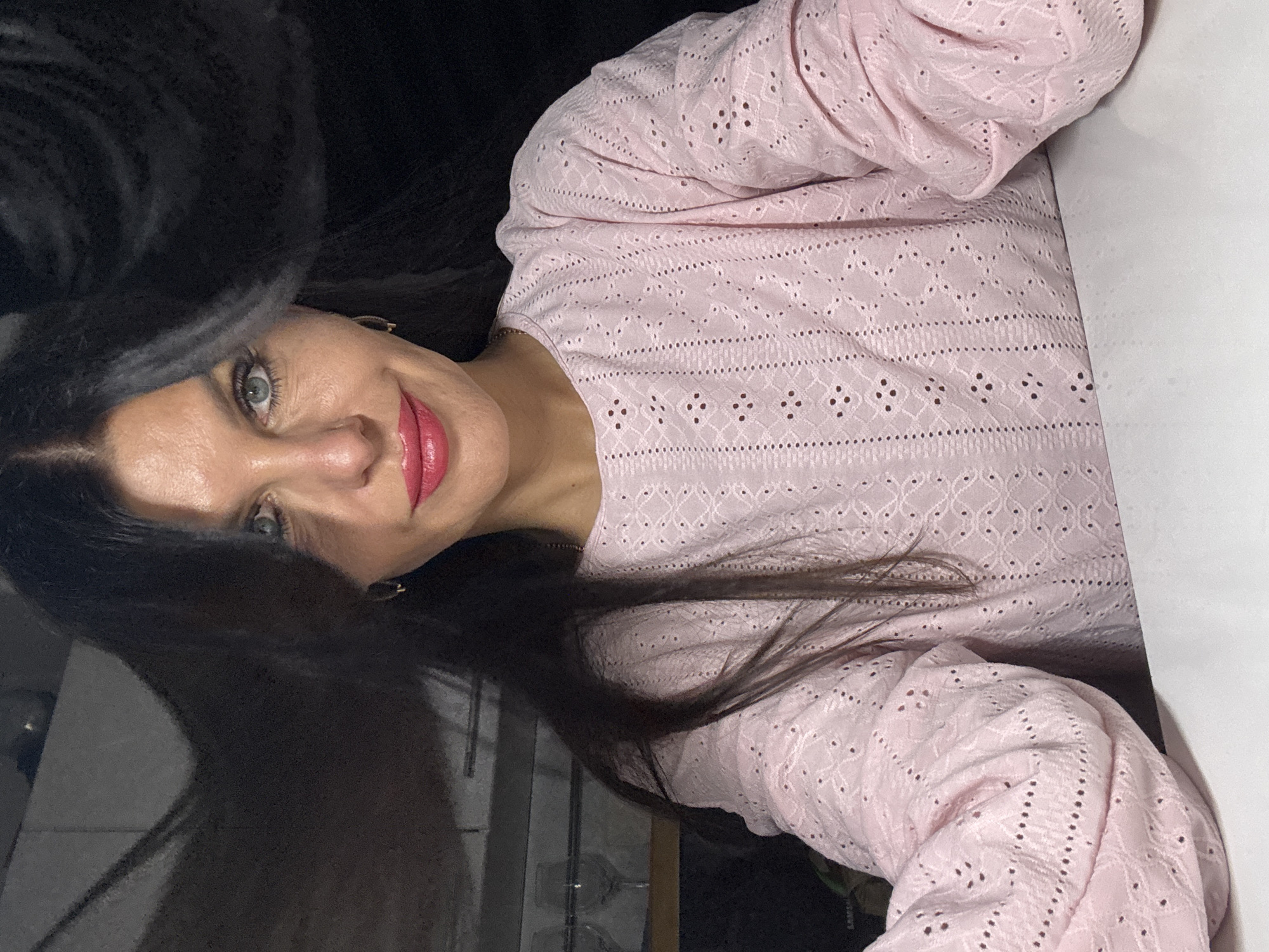 A woman with dark hair and light eyes is posing for a selfie. She is wearing a light pink, long-sleeved top with a patterned, eyelet-style design. She has red lipstick on and is smiling slightly. The background is dark and indistinct, with a hint of what may be a white surface on the right side of the frame and a glimpse of a window and wall on the left. There is insufficient information to ascertain the location of Chaikovsky, Russia.