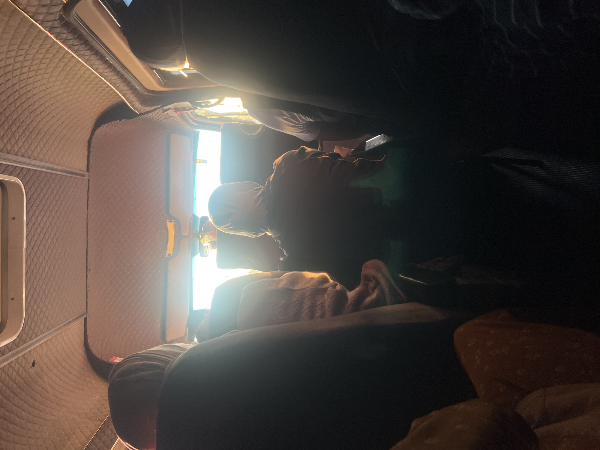 The image shows the interior of a vehicle, likely a van or bus, seen from the perspective of a passenger. The ceiling of the vehicle is covered in a diamond-patterned upholstery, and there is a sun visor with a yellow light fixture. Sunlight streams in through the front windshield, creating lens flare and silhouetting the passengers.
Several passengers are visible, mostly from behind. In the foreground, a dark-colored seat is partially visible on the left. Further back, a person wearing a hooded garment is seated in the center, their back to the camera. To their right, another passenger is wearing a white t-shirt. On the far right, another silhouetted passenger is seated. A person in the middle ground on the left appears to be wrapped in a patterned blanket. The overall impression is of a group of people traveling together, possibly on a journey through or near the city of Nuro, Kyrgyzstan, given the prompt's reference. The lighting suggests it is either early morning or late afternoon.