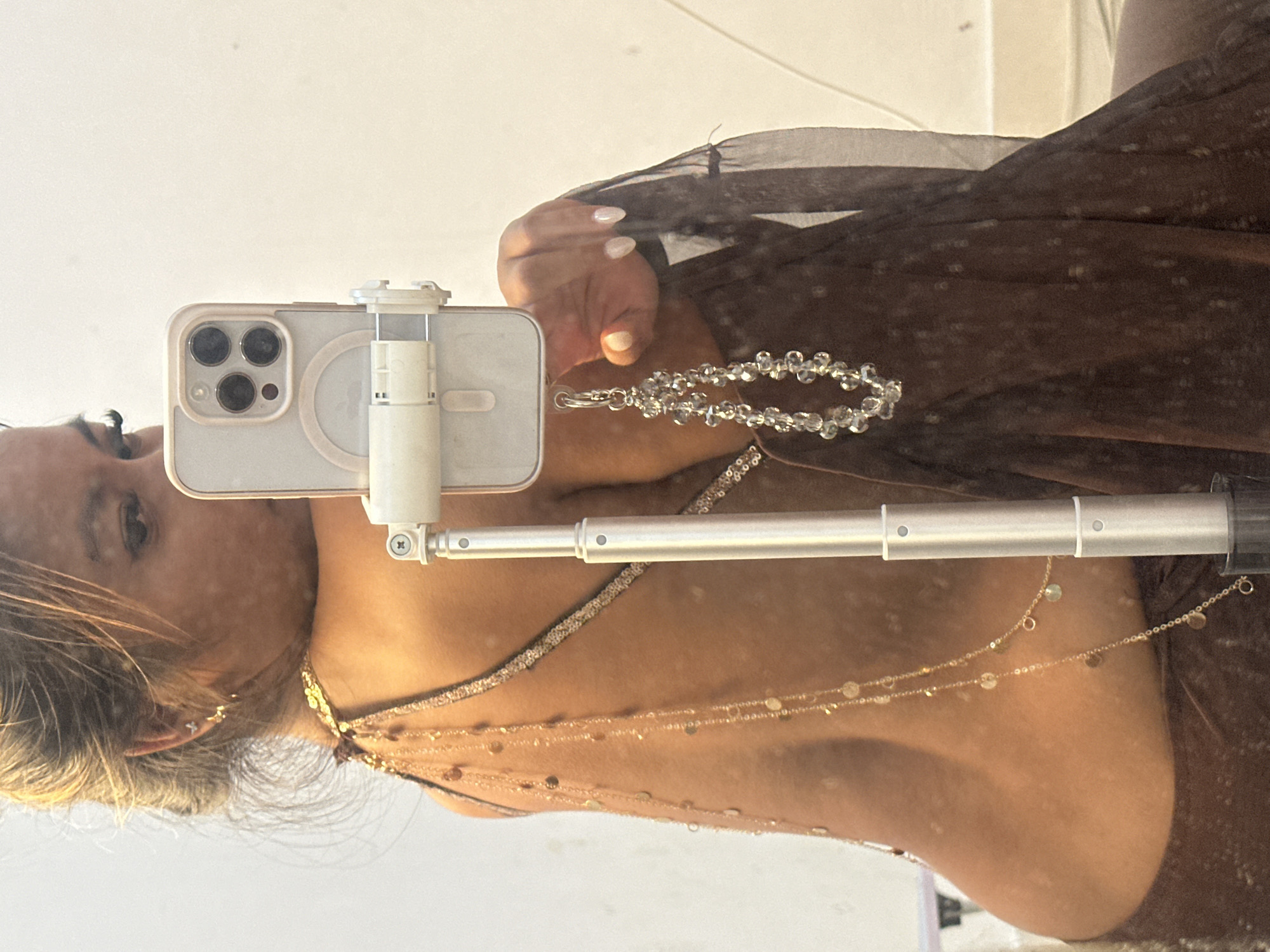 A selfie taken in a mirror shows a woman holding a phone attached to a selfie stick, positioned in front of her face. She is wearing a brown dress with layered gold necklaces. A beaded bracelet dangles from her hand. Her hair is blonde and pulled back. The phone is a white iPhone with three camera lenses. The background reflects a bright indoor space. The location is Celuk, Indonesia.