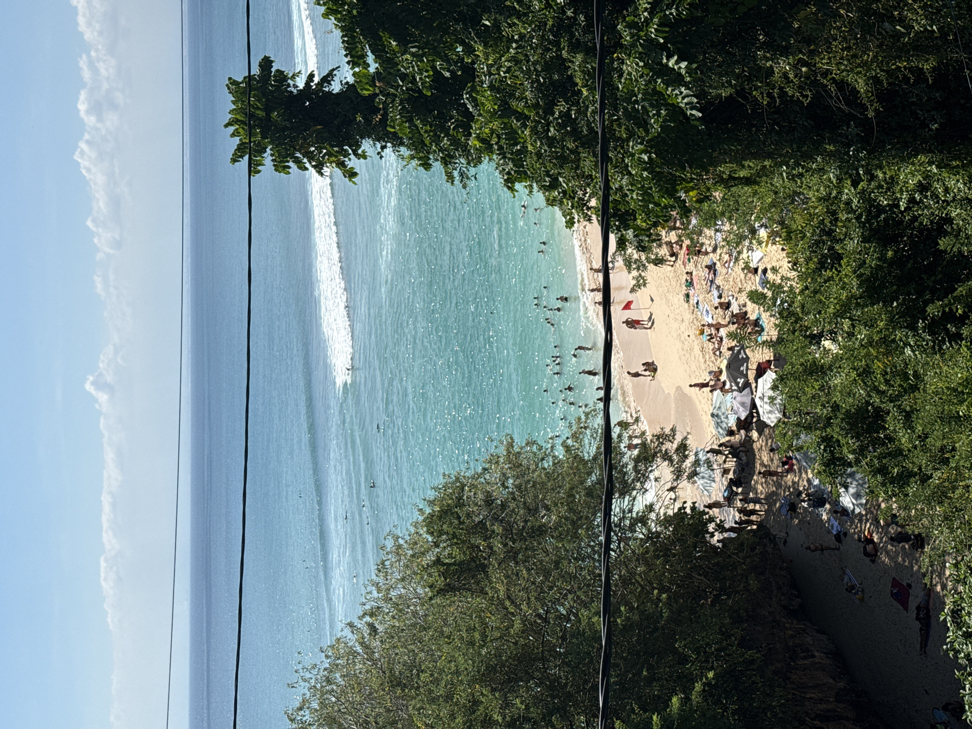The image captures an overhead view of a bustling tropical beach, likely in Kuta Selatan, Indonesia. The scene is dominated by the vast expanse of the ocean, transitioning from a deep blue horizon to sparkling turquoise waters closer to the shore, where white waves gently break.
Numerous people are visible enjoying the sunny day. Many are in the clear, inviting ocean, swimming or wading in the shallows. On the light-colored sandy beach, other individuals are scattered, some relaxing under colorful beach umbrellas or on towels, while others are seen walking or standing. Lush green foliage from dense trees and bushes frames the beach from the left, right, and foreground, indicating a natural, perhaps cliffside, setting overlooking the bay.
The time of day appears to be mid-day, characterized by bright, direct sunlight that illuminates the water's surface, creating shimmering reflections. The sky above is a clear, vibrant blue with a few fluffy white clouds visible in the distance. Two thin, dark wires, possibly power or communication lines, stretch horizontally across the foreground, slightly bisecting the view. A small red flag is noticeable on the beach, possibly signifying a lifeguard station or specific swimming conditions. No discernible text is visible within the frame. The overall atmosphere suggests a lively and popular beach destination.