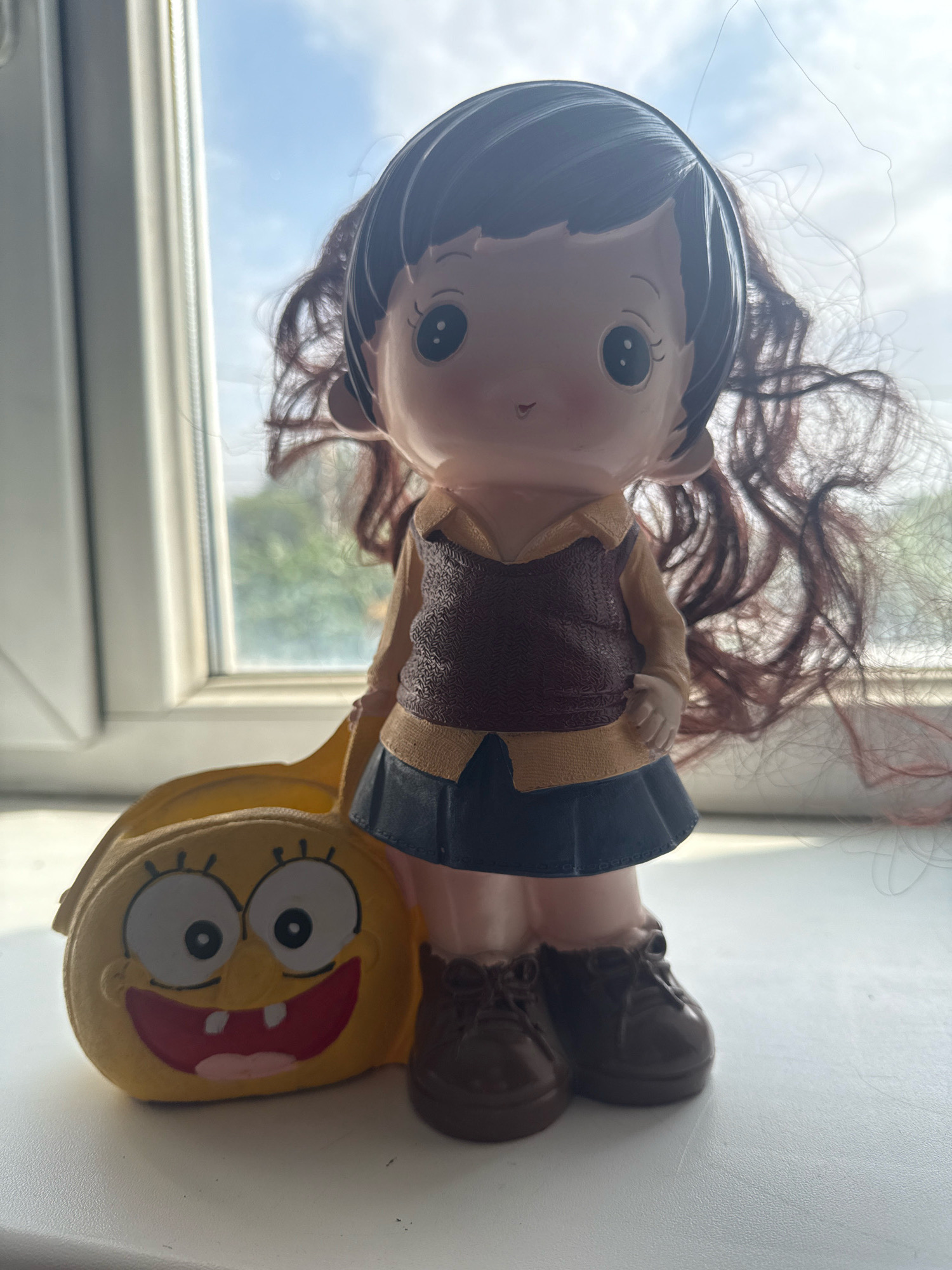 A doll with dark, curly hair stands next to a yellow, Spongebob-themed bag on a windowsill. The doll is wearing a collared shirt, a vest, a dark skirt, and brown shoes. The bag has Spongebob's face with wide eyes and a large grin. The background shows a window with a view of trees and the sky. The scene is lit by natural light, suggesting it is daytime. This image was taken in Bishkek City, Kyrgyzstan.