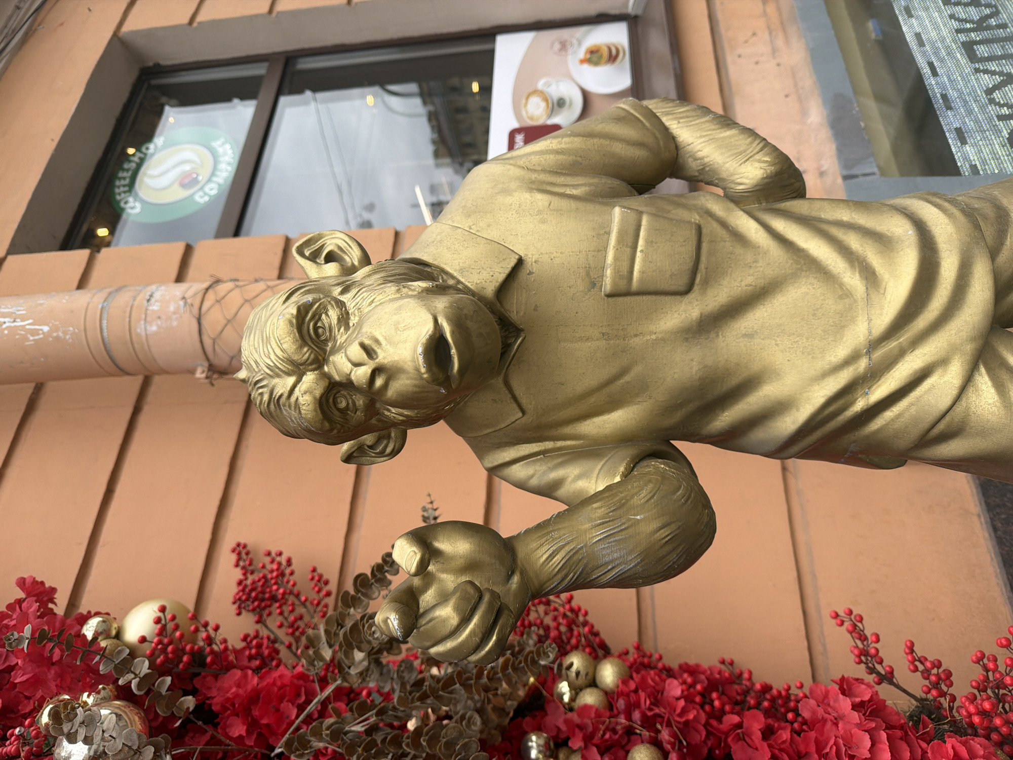 The image features a golden statue, possibly a monkey or ape, appearing to hang horizontally in front of a building facade. The statue is depicted wearing a jacket or shirt, with its mouth open and arms extended.
The immediate foreground and beneath the statue are adorned with festive red and gold decorations, suggesting a holiday or winter season. These decorations include clusters of bright red berries, red floral elements resembling poinsettias, and various shiny gold baubles, interspersed with some grey-green foliage.
In the background, a building exterior with orange or terracotta-colored panels is visible. Above this, a large window provides a glimpse into a establishment. On the window, a green circular logo is clearly visible, displaying the words "COFFEESHOP" and "COMPASS" in white text, along with a stylized graphic. Inside the window, a poster showcases images of food, such as pancakes, and coffee drinks, with some partially visible Cyrillic text, including "ВЫПИК".
The scene appears to be set during daytime, with reflections in the window indicating ambient light. The presence of the coffeeshop logo and the festive decorations suggest a commercial area. Given the Cyrillic text and the provided context, the location is Saint Petersburg, Russia.