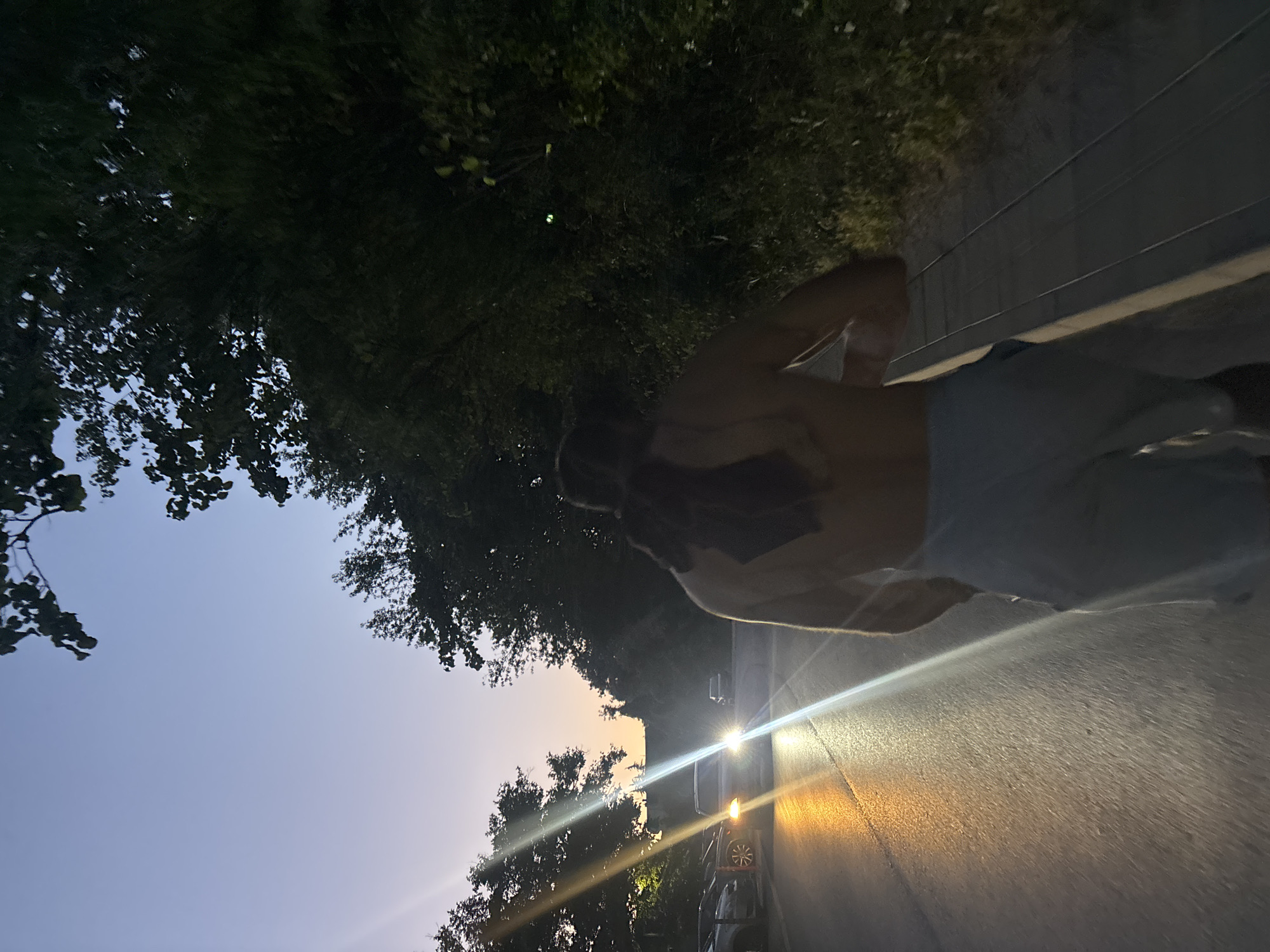 The image depicts a street scene at dusk, likely in Varna, Bulgaria, given the context of the request. In the foreground, a shirtless individual wearing light-colored pants is walking away from the camera, with a dark fabric draped over their shoulders. The person is partially blurred, suggesting movement. To the left, cars are parked along the side of a wet asphalt road, illuminated by streetlights and the headlights of an approaching vehicle. The road reflects the light, creating streaks of orange and white. Dense trees line the right side of the road, obscuring much of the background, and are silhouetted against a dusky sky that transitions from a pale blue to a soft orange near the horizon. The overall atmosphere is one of a transition from day to night in an urban or suburban setting.