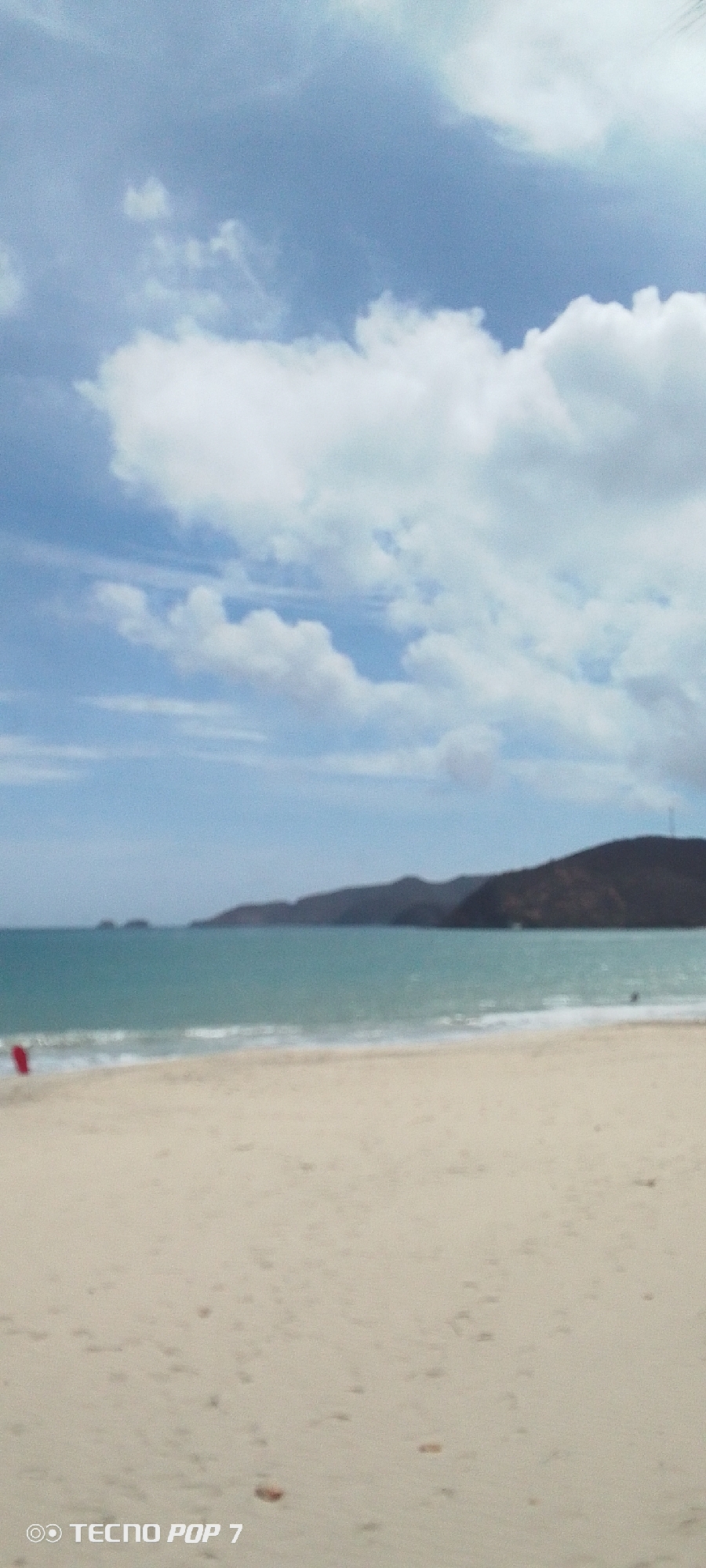 The image depicts a beach scene in Pedro González, Venezuela. The foreground shows a sandy beach with scattered small dark spots, possibly pebbles or debris. The middle ground features the turquoise ocean meeting the shore with small waves. In the distance, blurred, mountainous terrain is visible. The sky occupies the upper portion of the image, showcasing a bright blue hue with scattered white clouds. A person wearing a red item is present on the far left of the image. "TECNO POP 7" is seen at the bottom left.