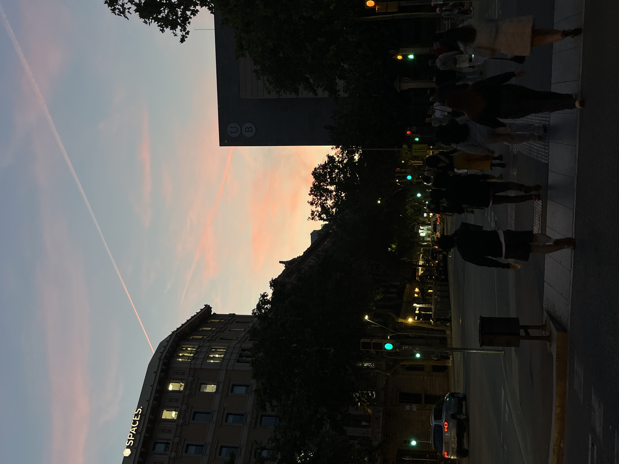 A nighttime street scene shows people crossing a crosswalk at a traffic intersection. The sky displays streaks of pink and grey, likely due to the sunset. Buildings are visible on the left side, including one with the word "SPACES" illuminated vertically. Traffic lights are seen along the street, with some displaying green lights. A street sign with the letter "U" and "B" is on a building. A car is stopped at the intersection. The city is Unknown, Unknown.