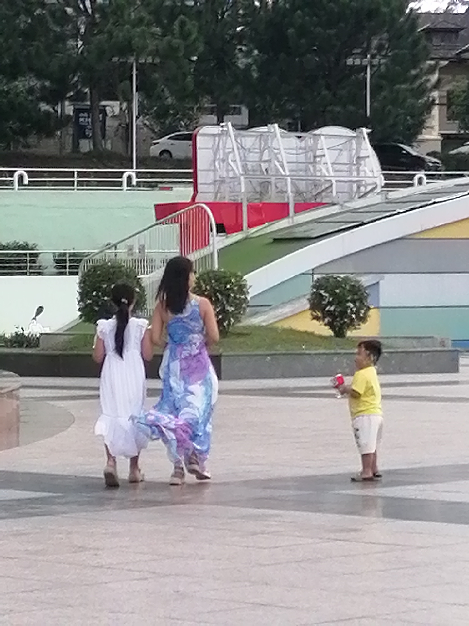In Da Lat, Vietnam, an outdoor scene unfolds with three individuals. A young boy, wearing a yellow t-shirt and white shorts, stands to the right, holding a red and white toy. To the left, two females are walking away from the camera. The female on the left wears a white dress, and the female on the right is in a flowing blue and purple dress. They are on a paved area, with trees and modern architecture in the background. A white railing with a red barrier is visible behind them, suggesting an elevated pathway or a recreational area. The environment appears to be a public space, possibly a park or a plaza in Da Lat.
