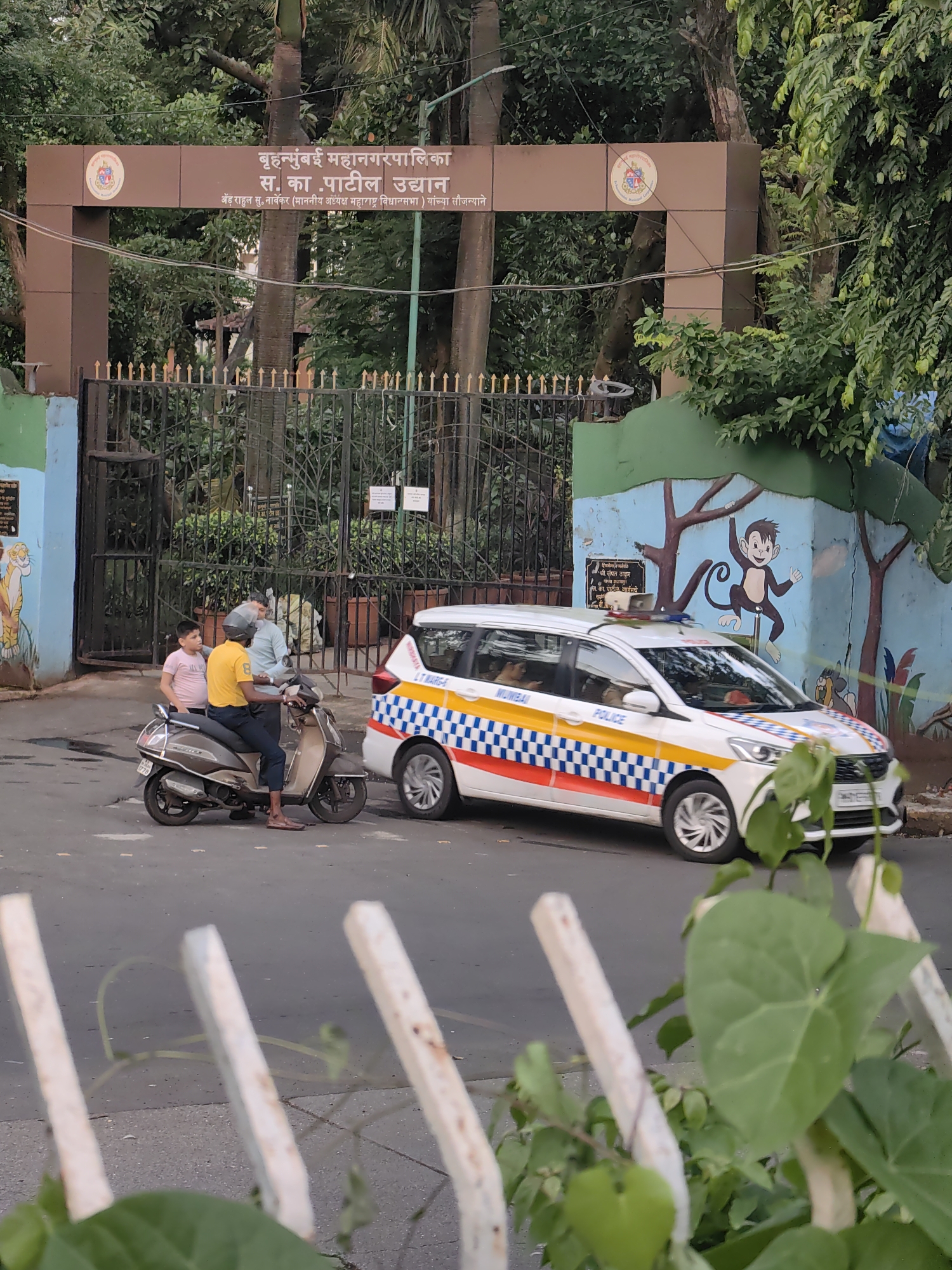 The image depicts the entrance to a park in Mumbai, India, indicated by the Marathi text on the signage above the gate, which translates to "Brihanmumbai Municipal Corporation S. K. Patil Garden." The gate itself is made of black wrought iron with gold accents. A white police van with distinctive blue and red checkered stripes and the word "POLICE" written on its side is parked to the right of the gate. In front of the gate, a man wearing a yellow shirt and dark pants is seated on a brown scooter, and a young boy in a pink t-shirt stands beside him. The background features lush green trees and a colorful mural on a wall to the right, depicting cartoon monkeys. The foreground is partially obscured by a white fence and some green foliage. The scene suggests a typical day at a public park in Mumbai, with a police presence noted.
