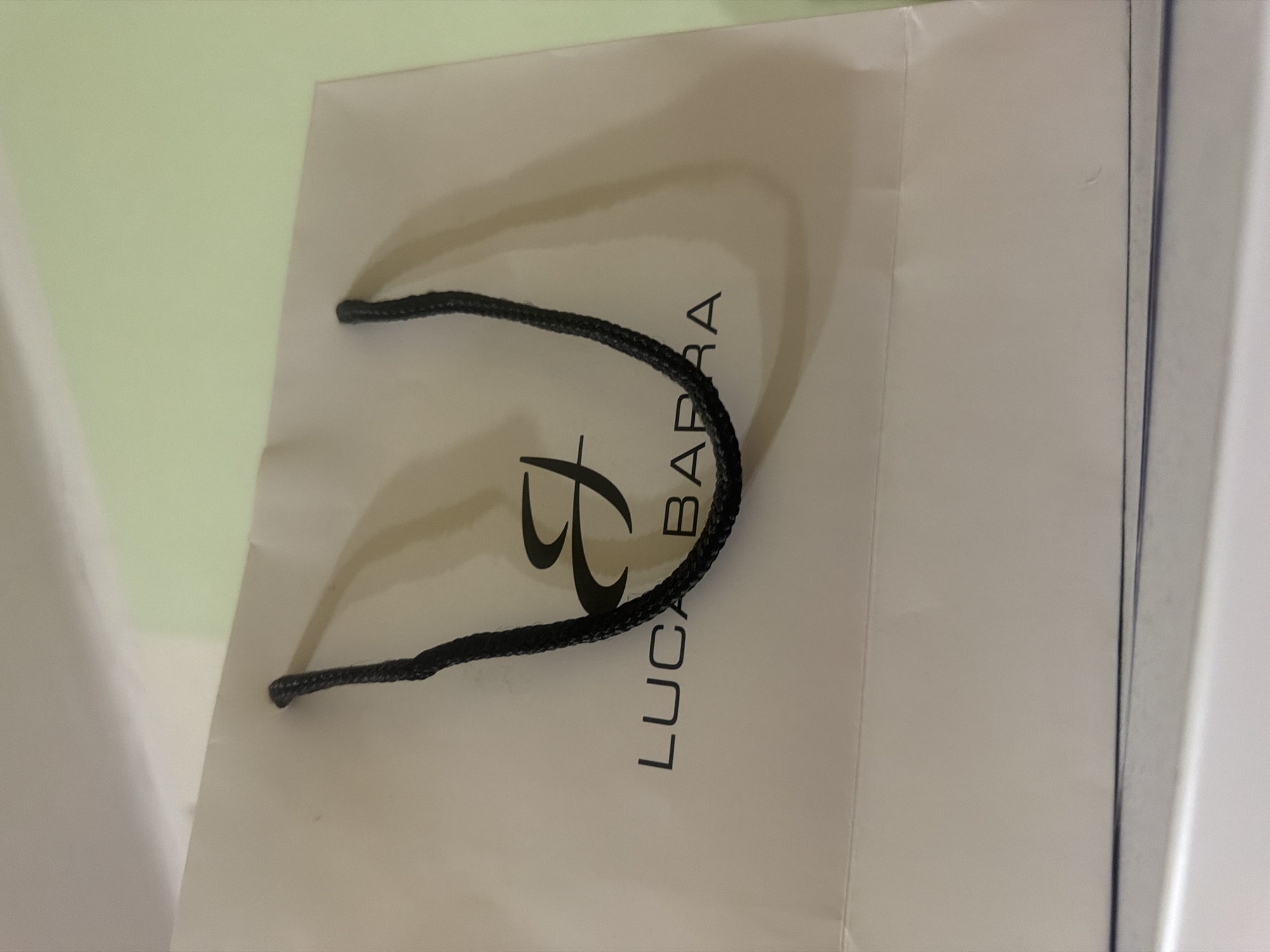 A close-up shot captures a partially visible white shopping bag. The brand name "LUCA BARRA" is vertically printed in black along with a stylized logo on the bag's surface. A black rope handle is visible, positioned over the brand name. The bag appears to be situated against a light green and white surface.