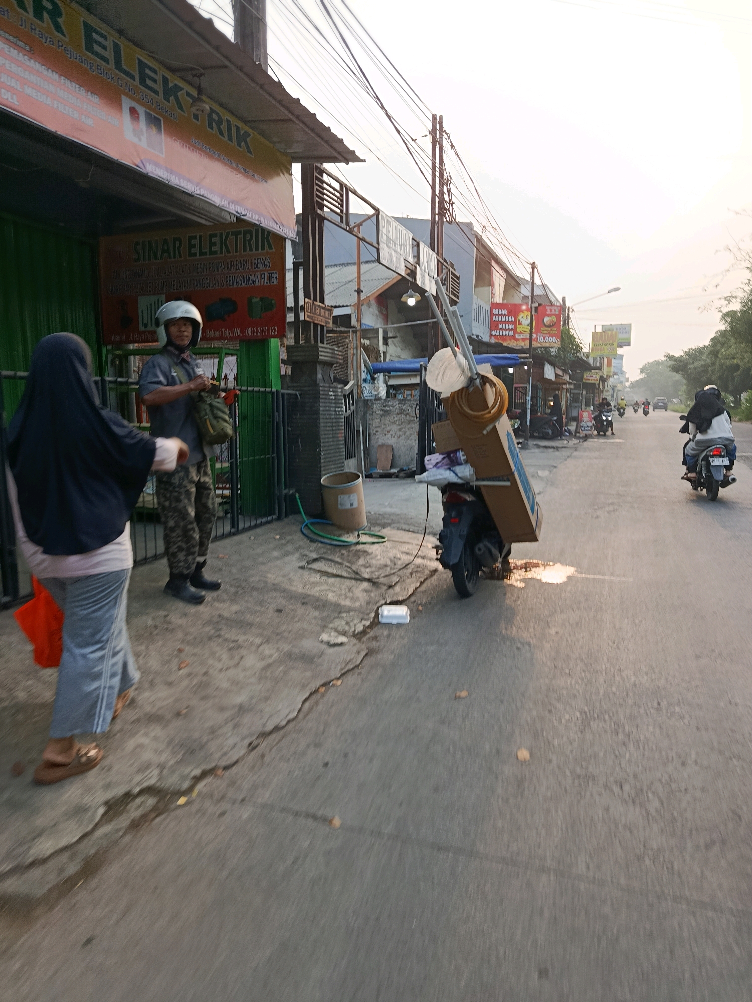 This is a medium-angle, eye-level, outdoor shot taken at street level in Bekasi, Indonesia. In the foreground, a person wearing a black hijab and a gray and pink outfit walks away from the camera, carrying an orange bag. To their left, a man in a gray short-sleeved shirt, camouflage pants, a white helmet, and black boots stands near a green metal gate. He is carrying a green bag over his shoulder. Behind them is a building with signage that reads "SINAR ELEKTRIK". A motorcycle is parked on the right side of the frame, loaded with a large cardboard box and a coiled hose. In the background, other motorcycles and a car are visible on the road, with more buildings lining the street. The sky is bright and hazy, suggesting it is either early morning or late afternoon. The ground is paved and appears wet in places, with some debris scattered around.
