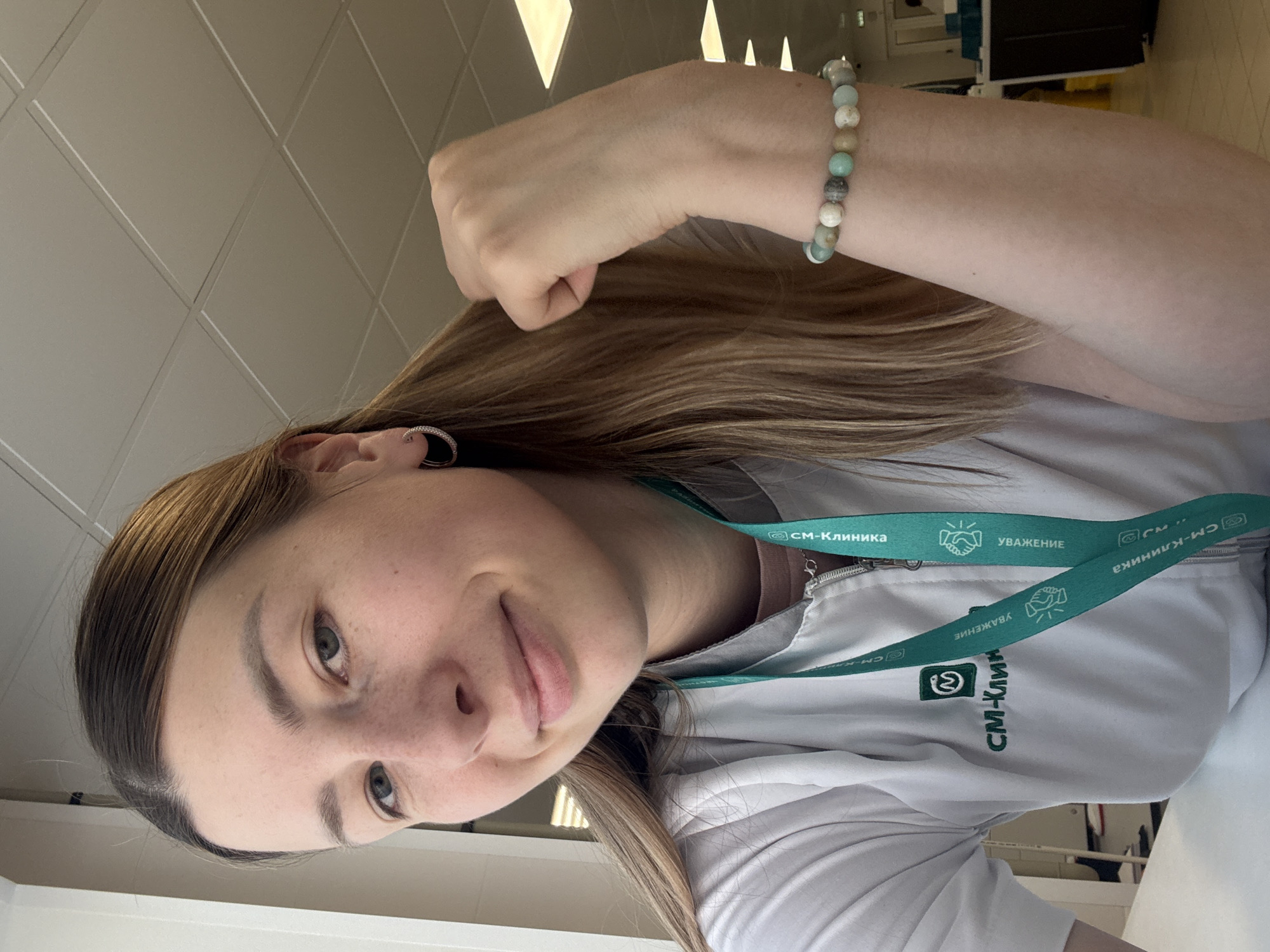 A woman is posing for a selfie. She is wearing a white top and a lanyard with the words "CM-Clinic уважение" (CM-Clinic Respect) written on it, which suggests that she works at the "CM-Clinic". She is wearing a beaded bracelet on her left wrist. The picture was taken indoors under artificial lighting. The ceiling has square panels. The picture may have been taken at the CM-Clinic, which has a location in the city of Khimki, Russia.