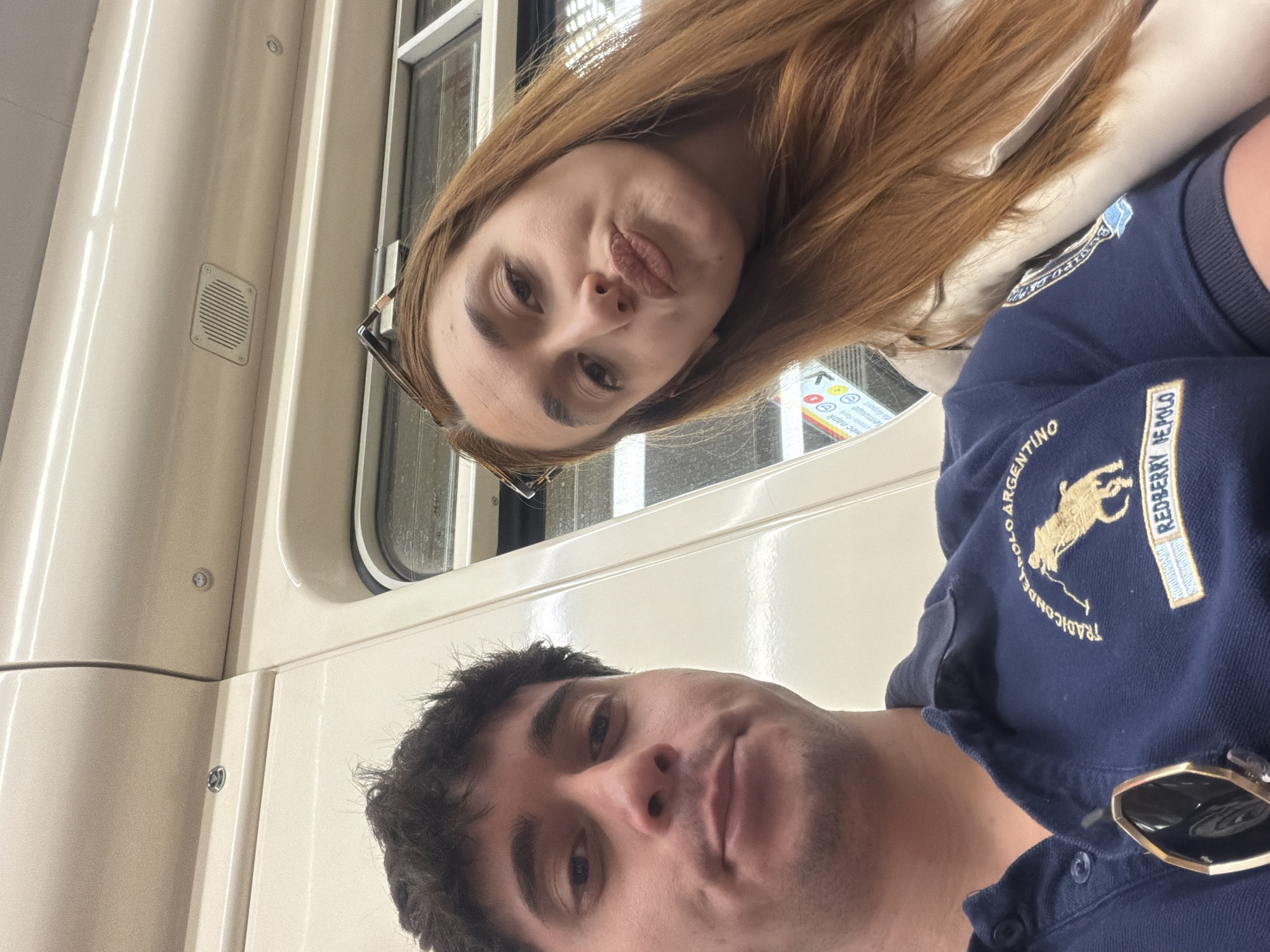 The image shows two people in what appears to be a train car. The interior of the train is off-white with a large window. A woman with long, auburn hair is positioned at the top of the frame, making a face towards the camera. She is wearing glasses. A man with dark, curly hair is positioned lower in the frame. He is wearing a dark blue polo shirt. The Polo shirt has sunglasses clipped to the collar.
There is nothing to show the location is Sofia, Bulgaria.