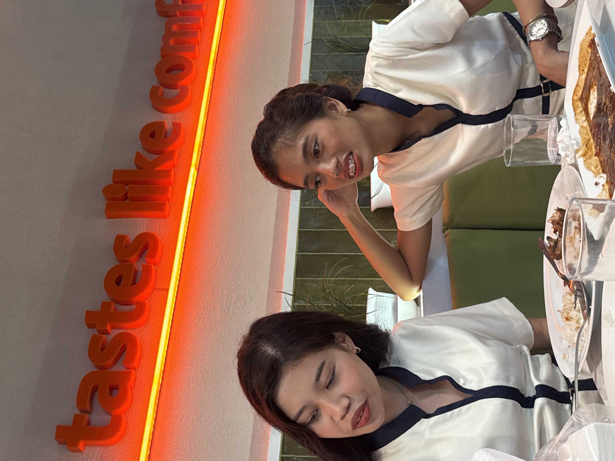 The image depicts two young women seated at a table in a restaurant in Davao City, Philippines. Above them, a neon sign with orange lettering reads "tastes like comf". The woman on the left has dark hair and is wearing a white shirt with a navy blue collar. She is looking down and to her left, with her mouth slightly open. The woman on the right has her hair tied up and is wearing a similar white shirt with a navy blue collar. She has braces on her teeth and is looking to her right, resting her arm on the back of the booth. Their clothing suggests they might be staff members. On the table, there are plates with food, cutlery, and glasses, indicating a meal in progress. The background shows green upholstery on the seating and some potted plants. The overall ambiance suggests a casual dining establishment.
