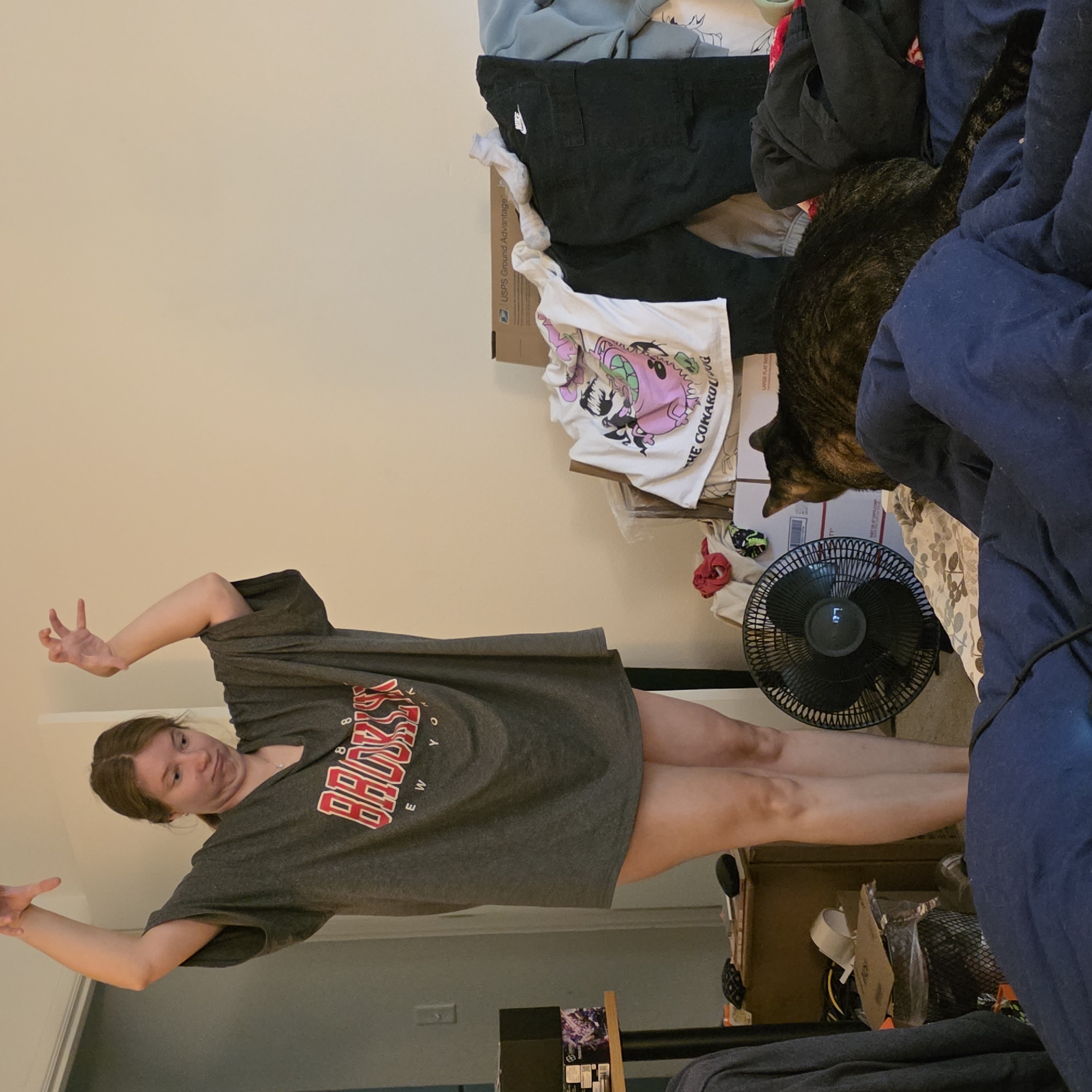In a cluttered indoor room, a young woman with brown hair pulled back stands with her arms raised in a playful gesture. She wears an oversized gray t-shirt with the word "BROOKLYN" in red and white lettering. Her facial expression is one of mock surprise or concentration. To her right, a tortoiseshell cat is perched on a pile of clothing, facing away from the camera. In the foreground, a dark blue comforter covers a bed, and a black oscillating fan is positioned to the left of the cat. Various items of clothing, boxes, and a trash can are scattered around the room, suggesting a disorganized living space. The scene appears to be set in a bedroom or dorm room. The lighting is artificial and casts soft shadows. The background consists of plain, light-colored walls. There are no visible indicators of the specific location within Portland, United States of America.