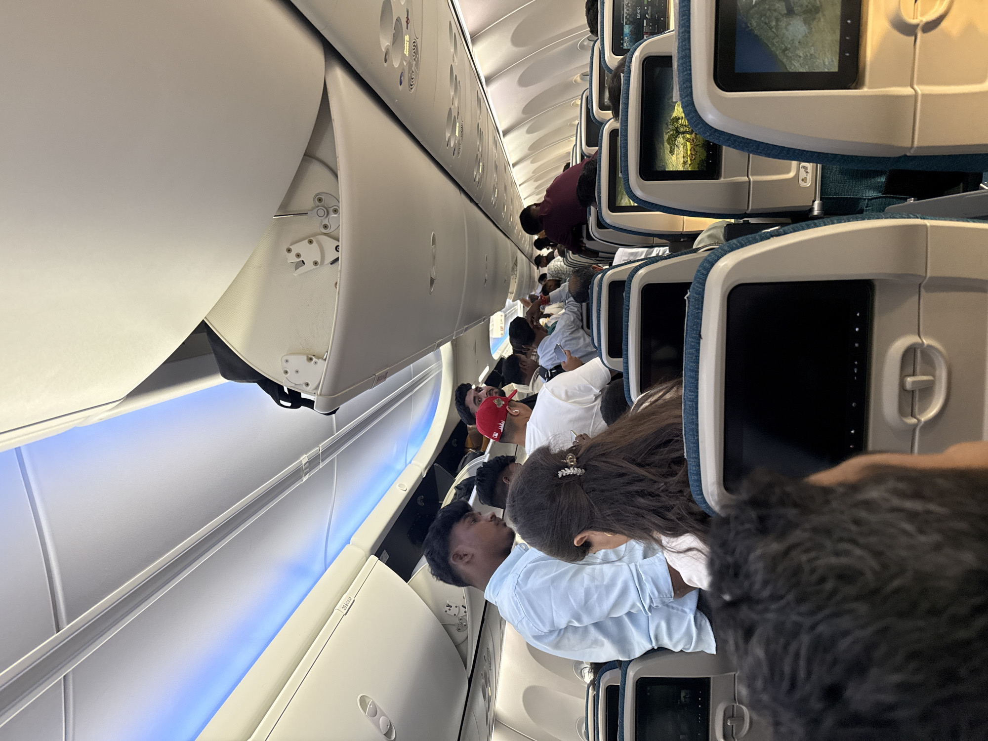 The image depicts the interior of an airplane cabin, likely en route from Dubai, United Arab Emirates. Numerous passengers are visible, some standing in the aisle and others seated. The overhead compartments are open, and the interior lighting is a soft blue. Seatback entertainment screens are visible, with one displaying a landscape scene. The overall impression is one of a crowded commercial flight.