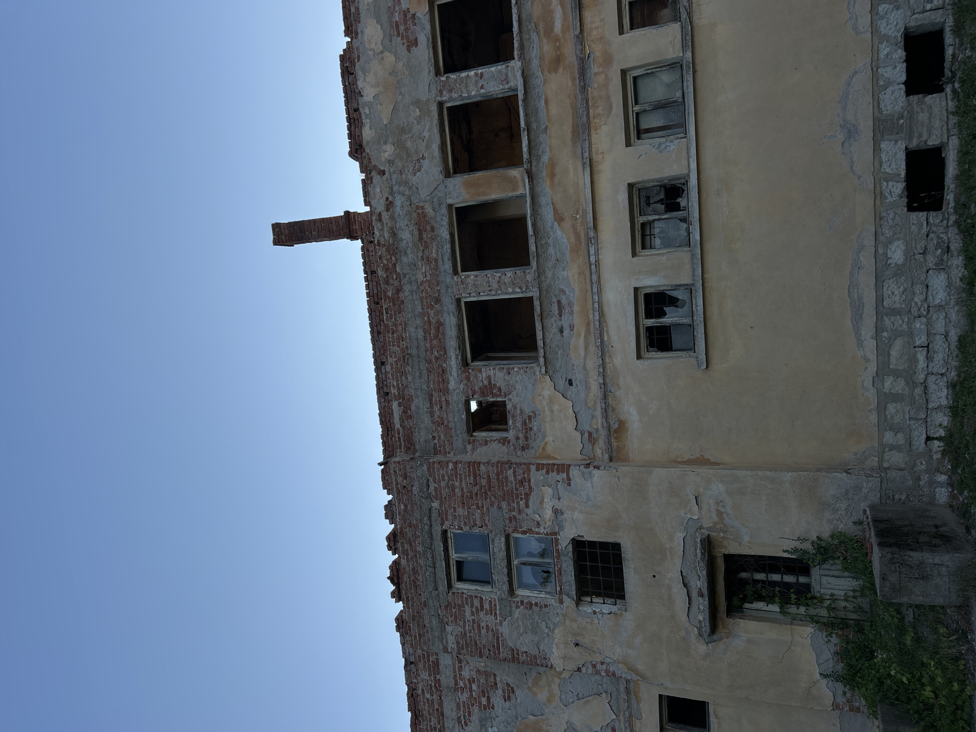 The image depicts a dilapidated, multi-story building with a brick and stucco facade. The building appears abandoned, with crumbling walls, exposed brickwork, and broken windows. A brick chimney stands on the roofline. Vegetation is growing around the base of the building, particularly near a doorway. The sky above is clear and blue. The building is located in the city of Belo pole, Bulgaria. There are no people or animals visible in the image, and no actions or interactions are taking place. The scene is static and conveys a sense of decay and neglect.