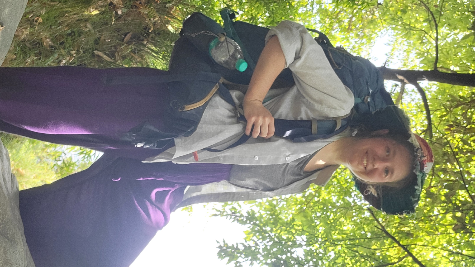 A young woman with fair skin and brown hair is smiling at the camera. She is wearing a light grey shirt, a dark grey undershirt, and dark purple pants. She has a large backpack on, with a blue water bottle attached. She is wearing a hat with colorful fabric. The background shows a forest with sunlight filtering through the leaves. The image has been rotated counter-clockwise. There is no information to verify that this location is Haini, India.