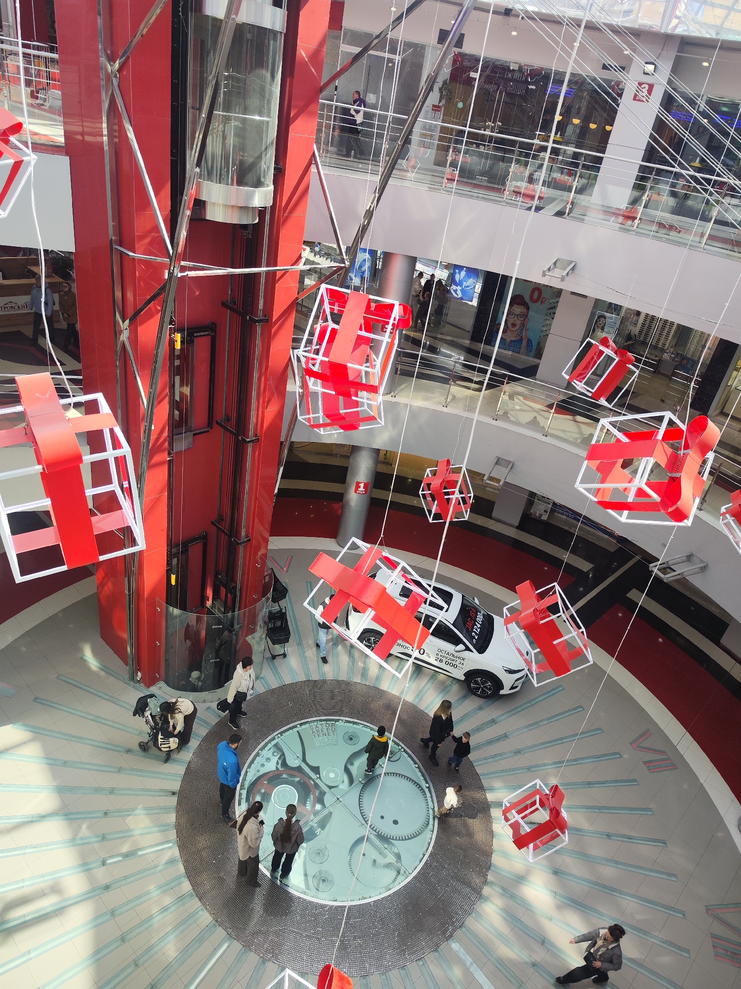 The image depicts an interior view of a shopping mall atrium. A large, central, red-colored elevator shaft with a glass-enclosed elevator is a prominent feature. Below the elevator is a circular floor design with a clock mechanism under glass. Several people are visible on the ground level, some standing near the clock, others walking around. A white JAC JS6 car is displayed, surrounded by several of the pedestrians. Suspended from the ceiling are numerous decorative white cube frames with red ribbon accents. Balconies of upper floors are visible around the atrium, with people present on some of them. Advertisements are posted on some of the upper floors. The shopping mall is located in Izhevsk, Russia.