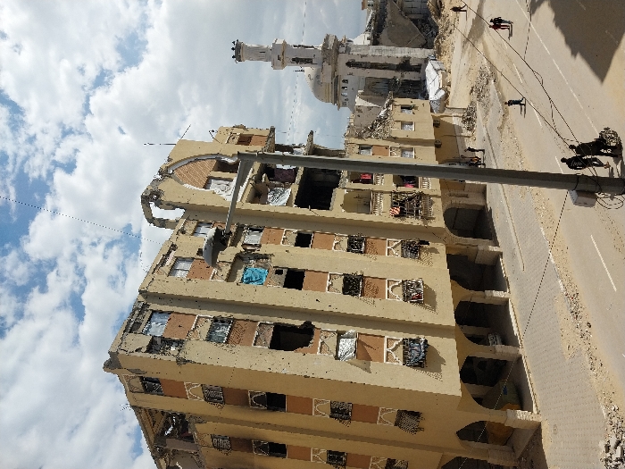 The image depicts a street scene in Khan Yunis, Palestinian Territory, characterized by significant structural damage. A multi-story residential building, approximately six floors high, dominates the left side, exhibiting extensive damage with large holes, exposed interiors, and broken window frames. Its beige facade is pockmarked, and the upper levels show severe structural compromise. Below, the building features arched ground-level supports, creating an open passage.
To the right, in the background, a mosque is visible, its white minaret partially collapsed at the top and its dome showing damage. Rubble and debris surround the mosque, with several white tent-like structures present nearby.
The asphalt street in the foreground is partially covered in dust and debris, with visible white lane markings. A grey utility pole with a streetlamp extends diagonally from the lower right into the upper middle, with wires traversing the frame; a bicycle leans against its base.
Several individuals are present on the street: two people are visible in the lower left, one pushing a wheelchair or cart. Another person walks alone in the mid-ground, and a pair of individuals walks further to the right. Additional figures are sparsely distributed in the distance near the mosque. The sky above is partly cloudy.
