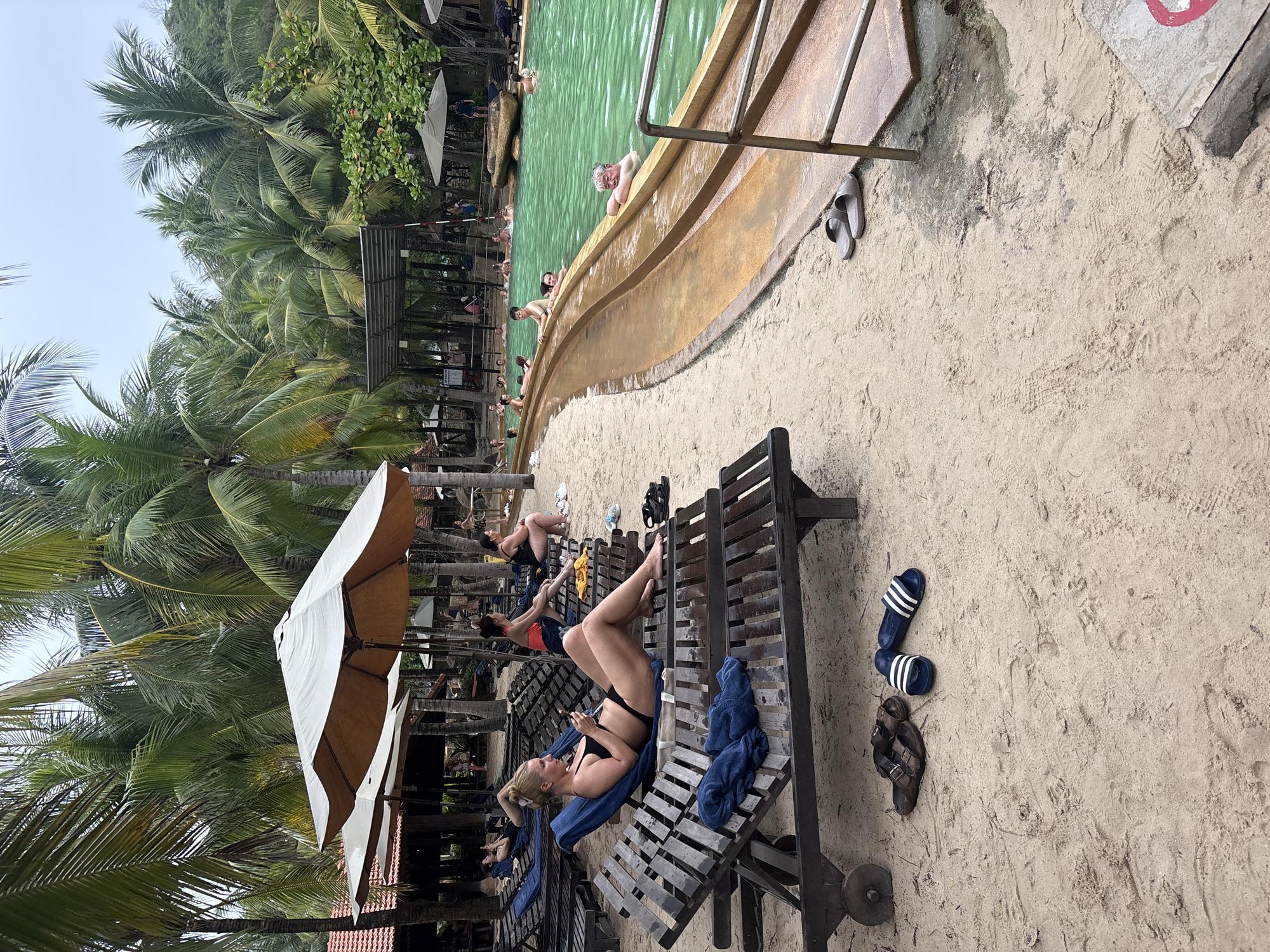 The image depicts a tropical resort pool area with sandy ground and numerous palm trees. In the foreground, a woman in a black bikini relaxes on a lounge chair, looking at her phone. Adjacent to her are two pairs of sandals on the sand, one brown leather and one blue and white striped. More lounge chairs and umbrellas are visible further back, with people sitting and reclining. To the right, a vibrant green swimming pool is occupied by several individuals, with a sloping water slide structure extending into it. Buildings with red-tiled roofs are partially visible behind the lounge chairs and palm trees. The overall atmosphere suggests a resort or hotel in a warm climate, likely Nha Trang, Vietnam, given the abundance of palm trees and the tropical setting.