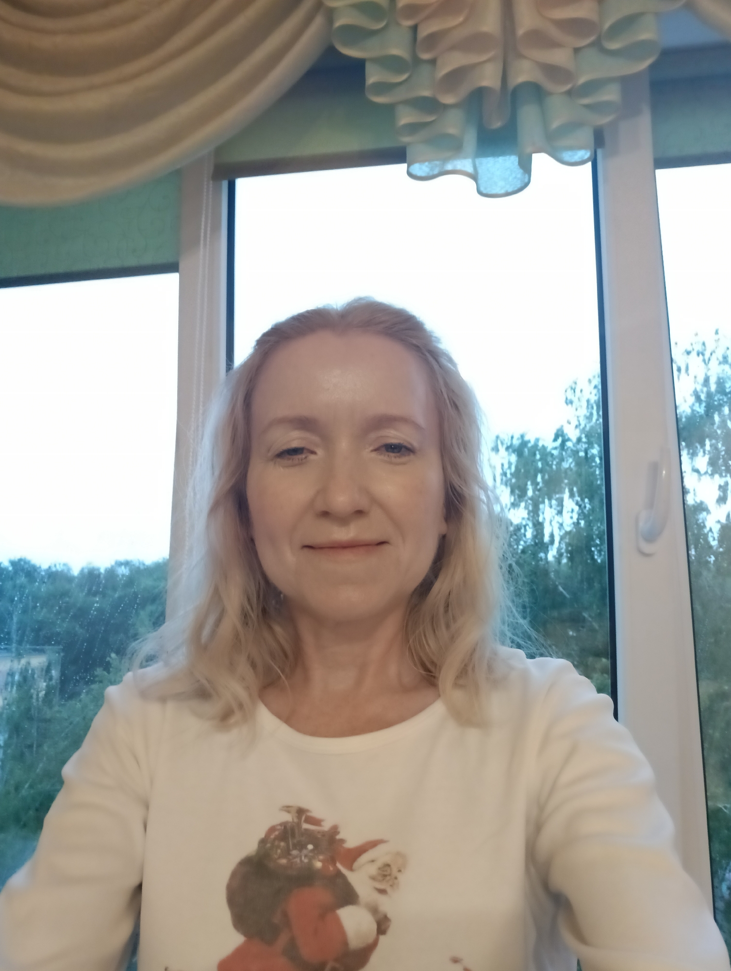A woman with blonde hair is shown in a medium shot, facing the camera. She is wearing a white long-sleeved shirt with a graphic of Santa Claus on it. The background appears to be a window with trees and sky visible outside. The top of the image shows a valance with a decorative, ruffled design. The overall scene suggests an indoor setting near a window, possibly a home. There is no specific indication of the city of Hrodna, Belarus.