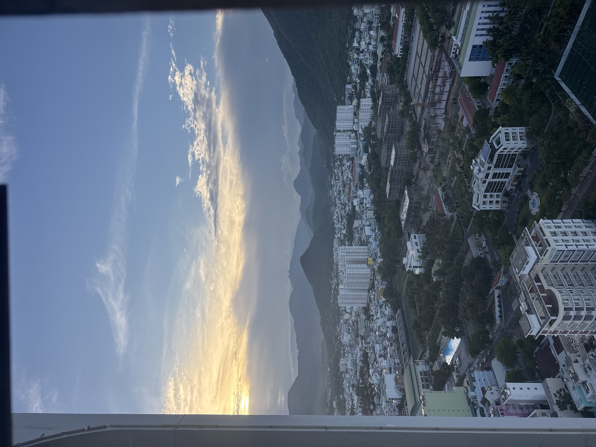 This is a vertical, aerial photograph taken from a high vantage point, looking out over a coastal city nestled against a backdrop of mountains. The sun is setting, casting a warm golden glow on the clouds in the sky to the left of the frame. The sky transitions from a bright blue to a hazy, pale yellow near the horizon, indicating the approaching sunset.
The city below is densely populated with a mix of buildings, including low-rise residential areas with white and colored roofs, as well as several taller, modern apartment buildings. Some buildings appear to be under construction, with scaffolding visible. Lush green trees are interspersed throughout the urban landscape, and a swimming pool with blue water is visible in the middle ground. In the distance, a series of majestic mountains rise, their peaks softened by the atmospheric haze of sunset.
The scene suggests a peaceful, evening atmosphere, with the natural beauty of the mountains and sky complementing the developed urban environment. The time of day is clearly late afternoon or early evening. There is no visible text in the image. The orientation of the photograph suggests it might have been taken from a high-rise building or a drone.