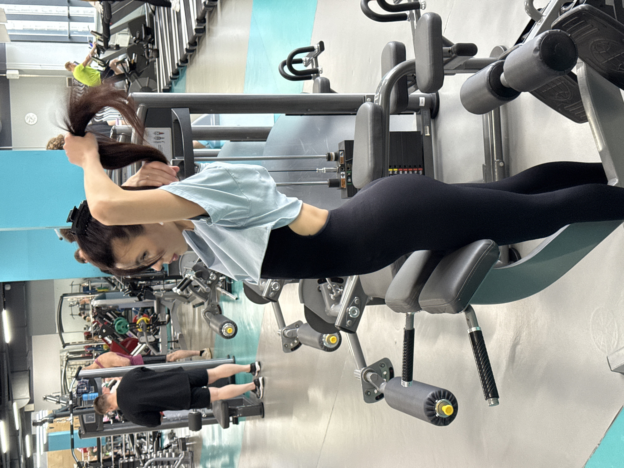 This photograph captures a dynamic scene within a modern gym located in Yekaterinburg, Russia. The primary subject is a woman in the foreground, positioned on a specialized gym machine, likely performing a back extension or hyperextension exercise. She is lying face down on the padded support, wearing black leggings and a light blue t-shirt which is slightly pulled up, revealing her midsection. Her long, dark hair is held up with both hands as she arches her back, demonstrating focused effort.
The gym environment is bustling, with several other individuals engaged in their workouts in the background. To the upper left, a man in a bright yellow t-shirt is visible on a cardio machine such as a treadmill or elliptical. Further in the background, another man in a black t-shirt and shorts is exercising on a different piece of equipment. Various fitness machines, including weight stacks, padded benches, and other training apparatus, fill the space, indicating a well-equipped facility.
The interior of the gym is brightly lit, suggesting it is daytime. The walls feature light colors, with distinct sections of light blue and pale green creating a contemporary aesthetic. No discernible text or specific branding is clearly visible on the equipment or walls. The overall atmosphere conveys a sense of activity and dedication to fitness.