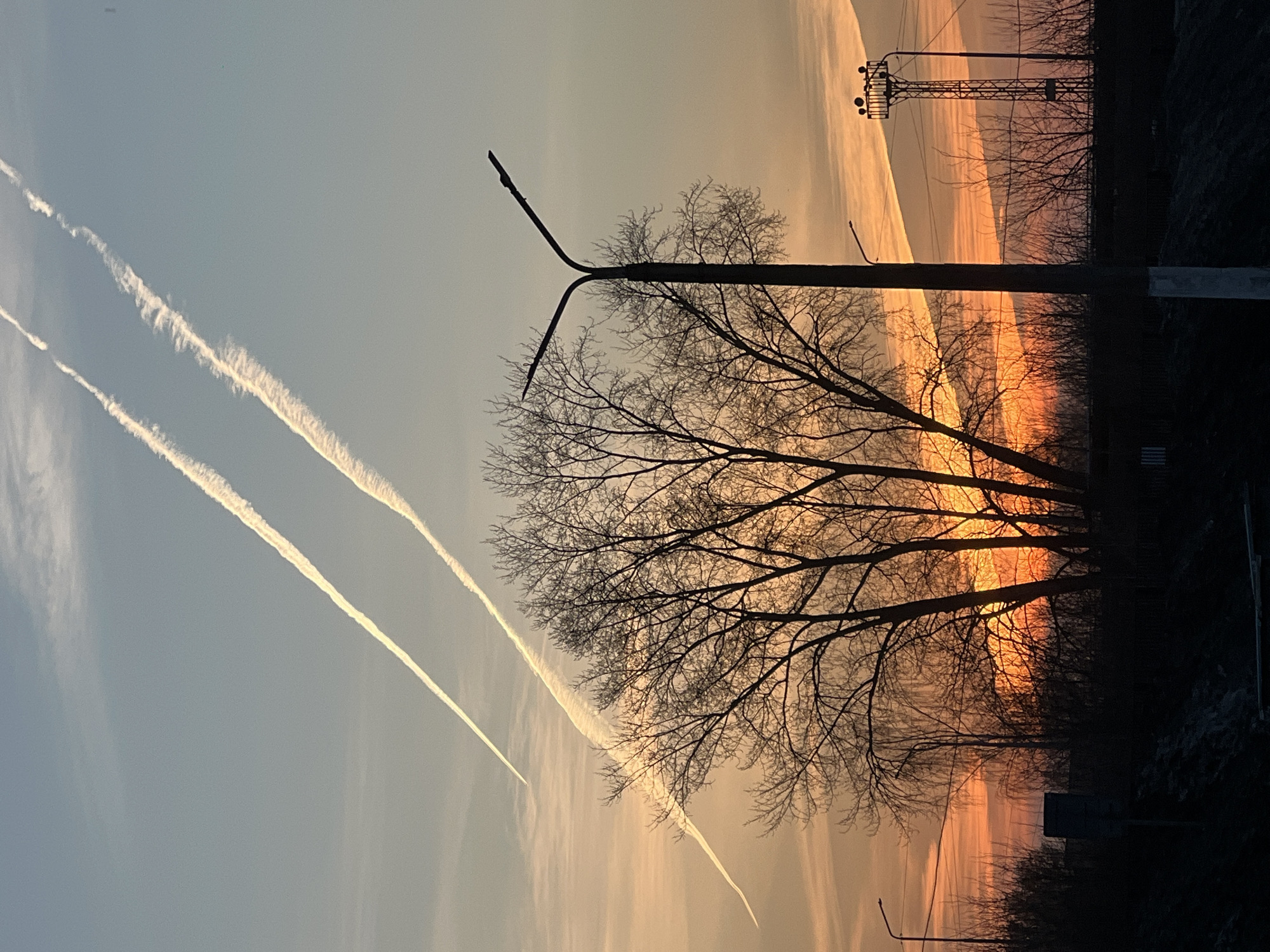 The image captures a sunset scene in Chaikovsky, Russia. In the foreground, a bare tree stands silhouetted against the warm orange glow of the setting sun, creating a stark contrast. A street lamp with a distinctive Y-shaped arm is also visible near the tree. In the sky, two distinct contrails, likely from passing aircraft, cut across the sky, adding a sense of depth and movement. In the background, a utility pole structure is visible against the backdrop of the fading light. The immediate foreground is dark, suggesting a low vantage point possibly at ground level. The overall atmosphere is one of tranquil beauty.