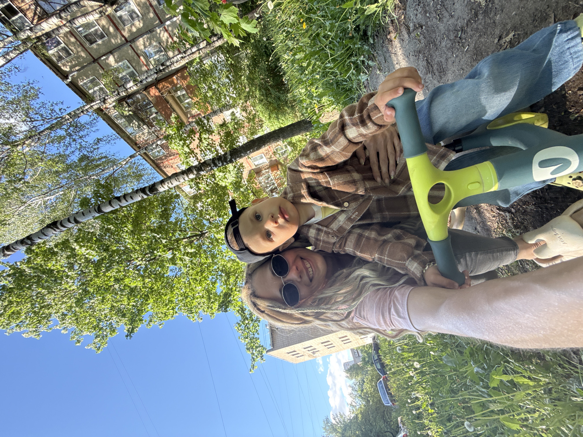 A low-angle shot shows a woman and a child on a balance bike outdoors. The woman has light-colored, braided hair, sunglasses, and a pink top. The child is wearing a brown plaid shirt and a black hat. They are both looking at the camera. The balance bike is green and yellow. Behind them are trees, grass, buildings, and a blue sky. The image was taken in Rusanovo, Russia.