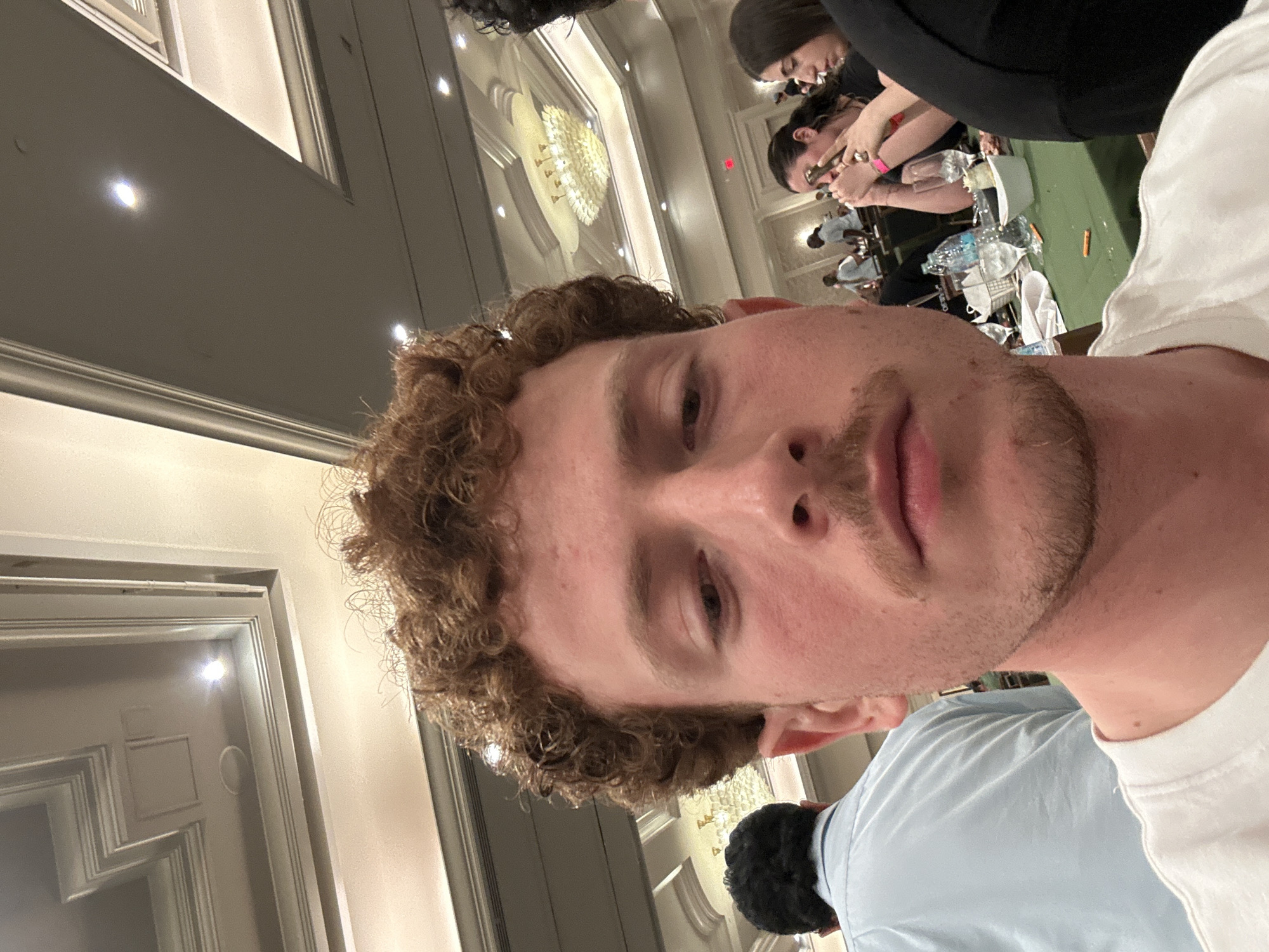 The image shows a close-up selfie of a young, light-skinned man with curly brown hair and a light beard. He is wearing a white T-shirt. Behind him and to the left is a gray wall with a decorative doorframe. In the background, other people are visible at tables, suggesting a banquet or event setting. There is a chandelier visible in the background. While the image may have been taken in Aventura, United States of America, there is not enough information available to confirm.