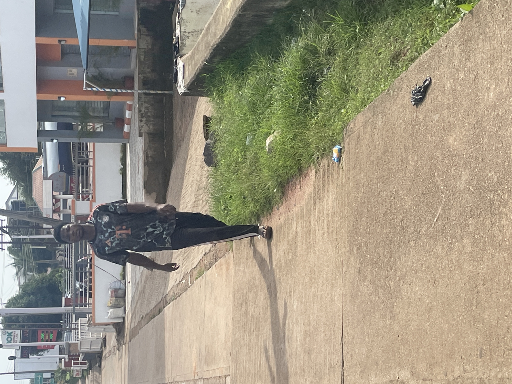 A young man walks on a concrete sidewalk next to a grassy area. He is wearing a dark t-shirt with a camouflage-like pattern and white stripes down the sides of his pants. In the background, there are buildings, some with orange accents, and trees. The scene appears to be a street in Gura Top, Nigeria. There are some discarded items on the ground near the grassy area, including a blue and yellow wrapper and a dark, tangled object. The lighting suggests it is daytime.