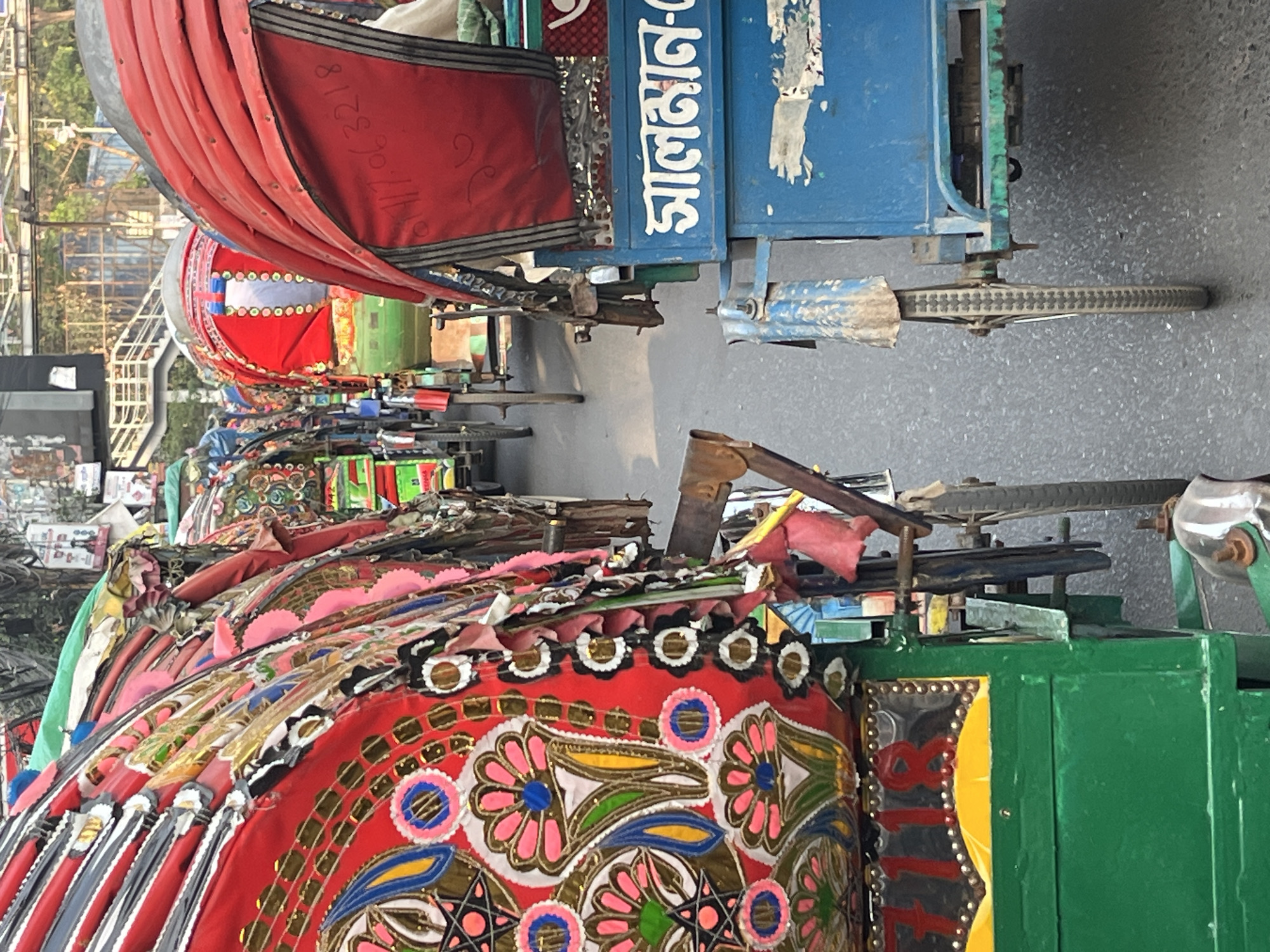 The image shows a row of parked cycle rickshaws in Dhaka, Bangladesh. The rickshaws are brightly decorated with colorful paint, fabric, and metallic accents. The seating area of each rickshaw is covered in red fabric, while the canopies above are decorated with floral and geometric patterns. In the background, there are buildings, trees, and a bridge or elevated structure. The pavement is gray and slightly worn. The photo is taken from an elevated perspective, looking down at the row of rickshaws. The quality of the image suggests it was taken with a mobile device.
