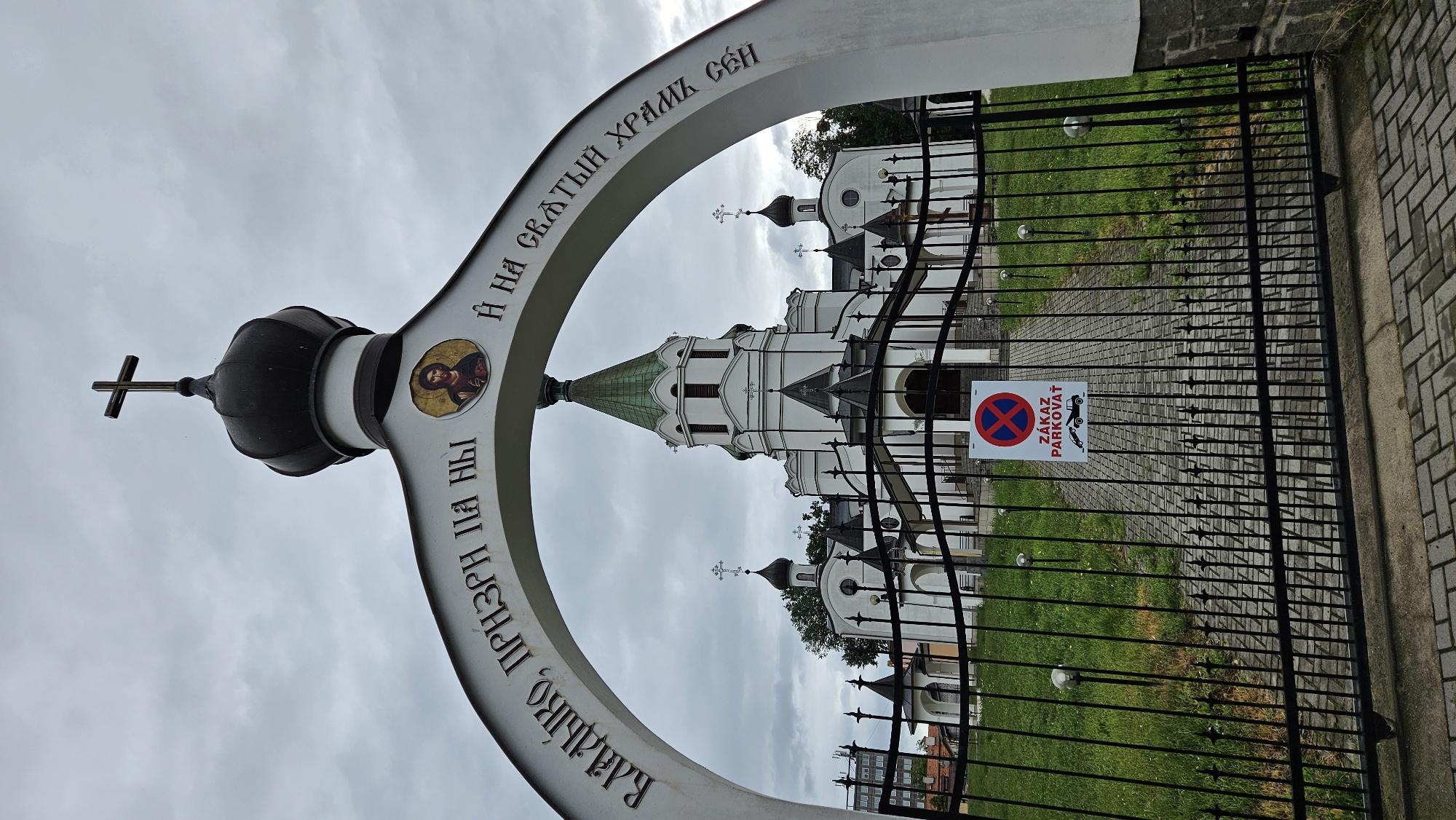 The image shows the entrance gate to the Orthodox Church of the Holy Cross in Prešov, Slovakia, on a cloudy day. The gate is made of black wrought iron and is closed. A sign with a red circle and a diagonal red line over a car icon is attached to the gate, indicating "No Parking" in Slovak. Beyond the gate, a paved path leads to the white church building, which features multiple domes and crosses. An archway above the gate has inscriptions in Cyrillic. The surrounding area is covered with green grass and trees.