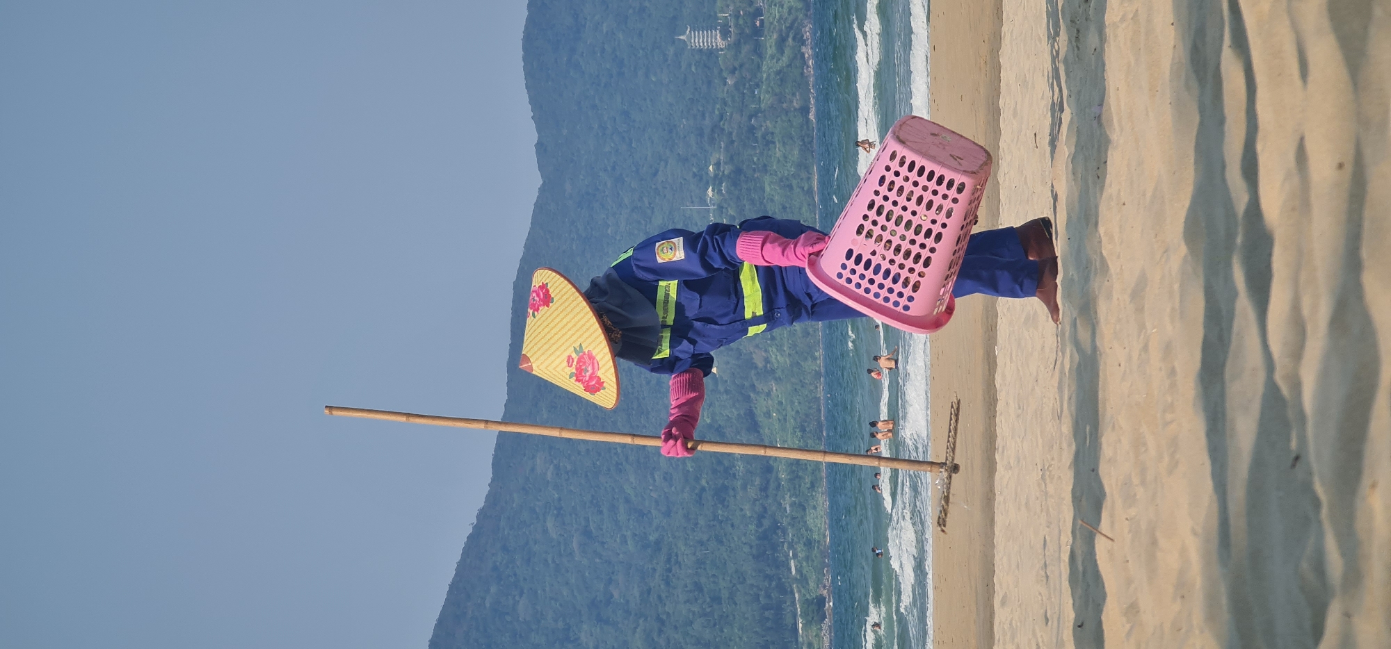 This image captures a person cleaning a sandy beach in Đà Nẵng, Vietnam, under a clear, bright blue sky. The individual is dressed in a blue uniform with reflective yellow-green strips and bright pink gloves. They wear a traditional Vietnamese conical hat (nón lá) decorated with red floral patterns.
The person is actively engaged in cleaning, holding a long bamboo pole or stick that extends towards the sand, and a pink, perforated plastic basket. Their posture suggests they are collecting refuse. One visible foot is bare, indicating they are walking directly on the sand.
In the background, the ocean displays turquoise waters with white waves breaking on the shore. Several people are visible swimming or wading in the sea. Beyond the coastline, towering green mountains rise prominently, and a distinctive multi-tiered structure, resembling a pagoda or temple, can be seen on a distant mountain peak, characteristic of the Son Tra Peninsula in Đà Nẵng.
The bright daylight casts clear shadows on the rippled sand, suggesting a time between late morning and early afternoon. The scene conveys a sense of environmental maintenance in a popular coastal setting. A circular emblem with text and a graphic, likely a uniform badge identifying their service, is visible on the person's left shoulder.