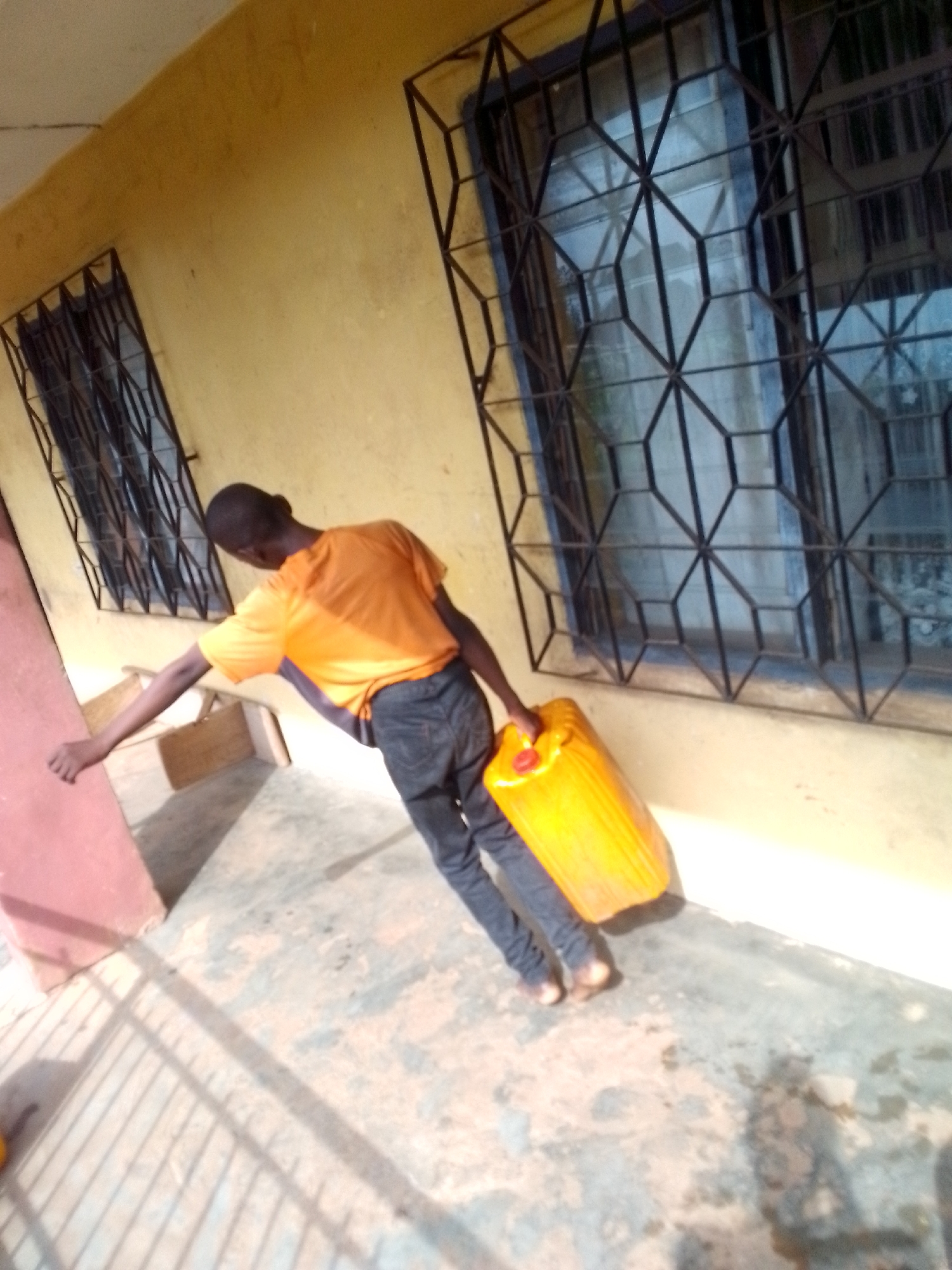A person with a dark complexion, wearing an orange t-shirt and dark jeans, is carrying a yellow jerrycan in their right hand. They are standing barefoot on a concrete surface beside a yellow building with two windows covered by geometric metal grilles. The location appears to be an outdoor space with shadows from a metal fence visible on the concrete ground. The city and specific location are not identifiable from the image.