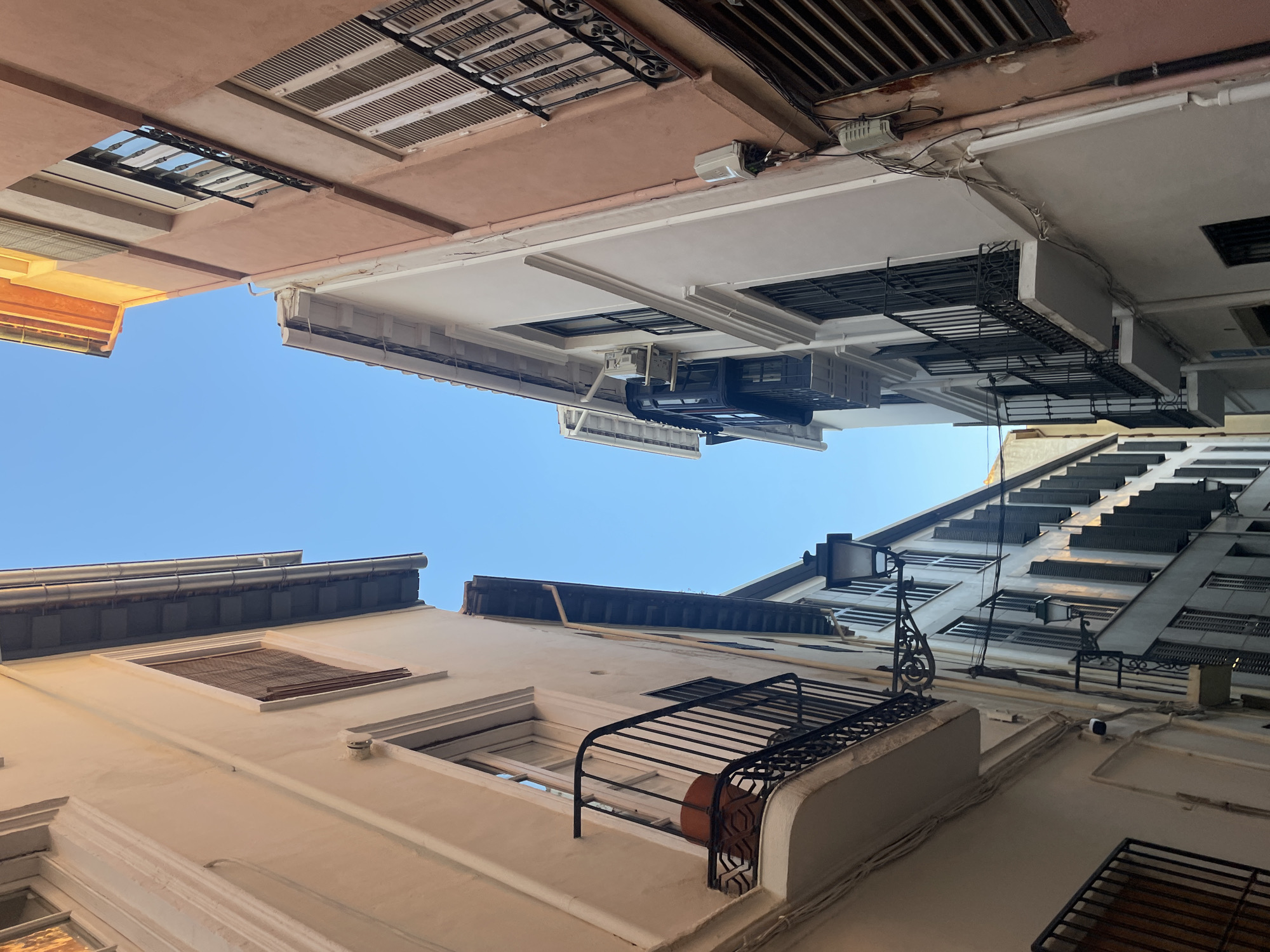 This image captures a narrow street in Granada, Spain, with tall buildings lining both sides and a clear blue sky overhead. The architecture is typical of the region, with balconies, shutters, and pastel-colored facades. The perspective is looking upwards, emphasizing the height of the buildings and the confined nature of the street. No people are visible in the scene. The street appears quiet and devoid of activity, bathed in natural light that casts subtle shadows. The focus is on the urban environment and architectural details, suggesting a residential or commercial area within the city.