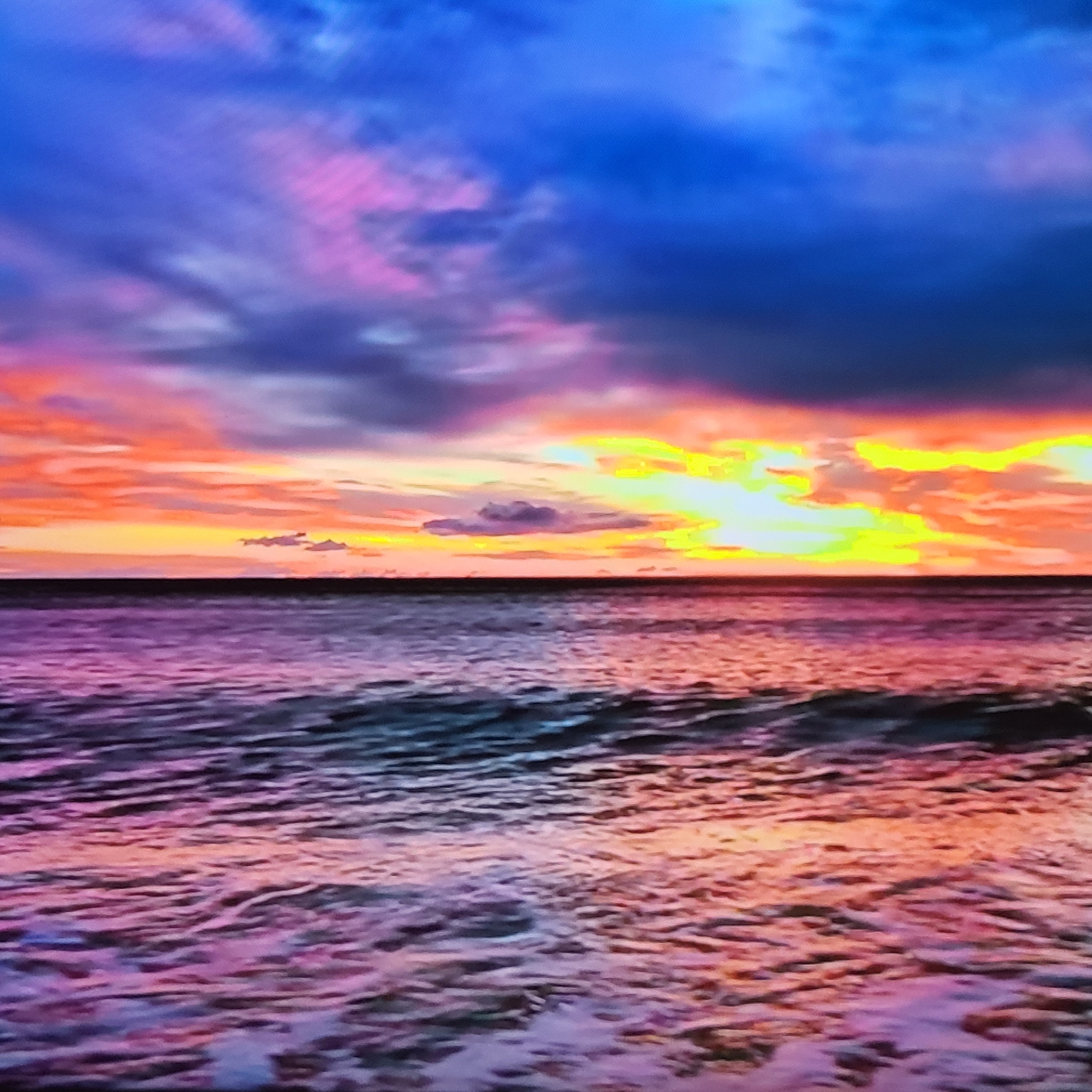 The image depicts a seascape at sunset. The sky transitions from a dark blue at the top to hues of pink, orange, and yellow near the horizon. The horizon line is a dark, almost black, strip that separates the sky from the water. The water reflects the colors of the sky, predominantly showing shades of pink, purple, and orange. The surface of the water is rippled with waves.
There is no information in the image to suggest that it is related to the city of Yerevan, Armenia.