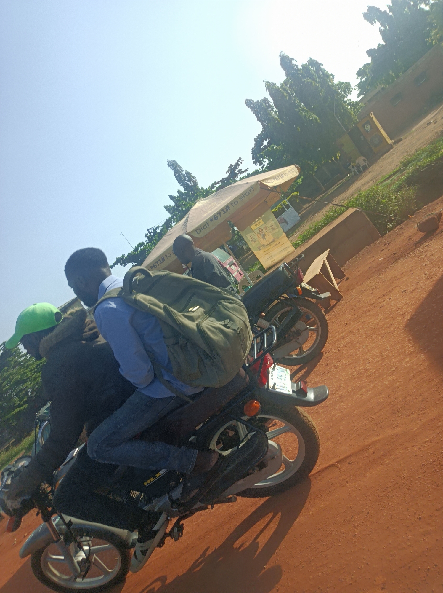 Three men are captured on two motorcycles, positioned on a reddish-brown dirt road under a bright, clear sky. In the foreground, a man wearing a blue shirt, jeans, and a large olive-green backpack is seated on a motorcycle. Another man with a bright green cap and a dark jacket is riding in front of him on the same motorcycle. Further back, a third man in a dark shirt is on a separate motorcycle, partially obscured.
The scene appears to be taking place during the day, likely mid-morning or early afternoon, given the strong sunlight and distinct shadows cast on the dusty ground. The weather is clear and sunny.
In the background, tall, dense green trees line the road, with a light brown building visible beyond them. A prominent beige umbrella structure, likely a roadside stall or service point, stands in the middle ground.
Visible text on the umbrella advertises "Dial *671# to start," suggesting a mobile network service code common in Nigeria. On the side panel of the motorcycle in the foreground, faint text "HLXC" and "ABEL" can be seen. A partial view of a license plate is visible on the rear of the motorcycle. The overall setting suggests a rural or semi-urban environment in Barnawa, Nigeria.