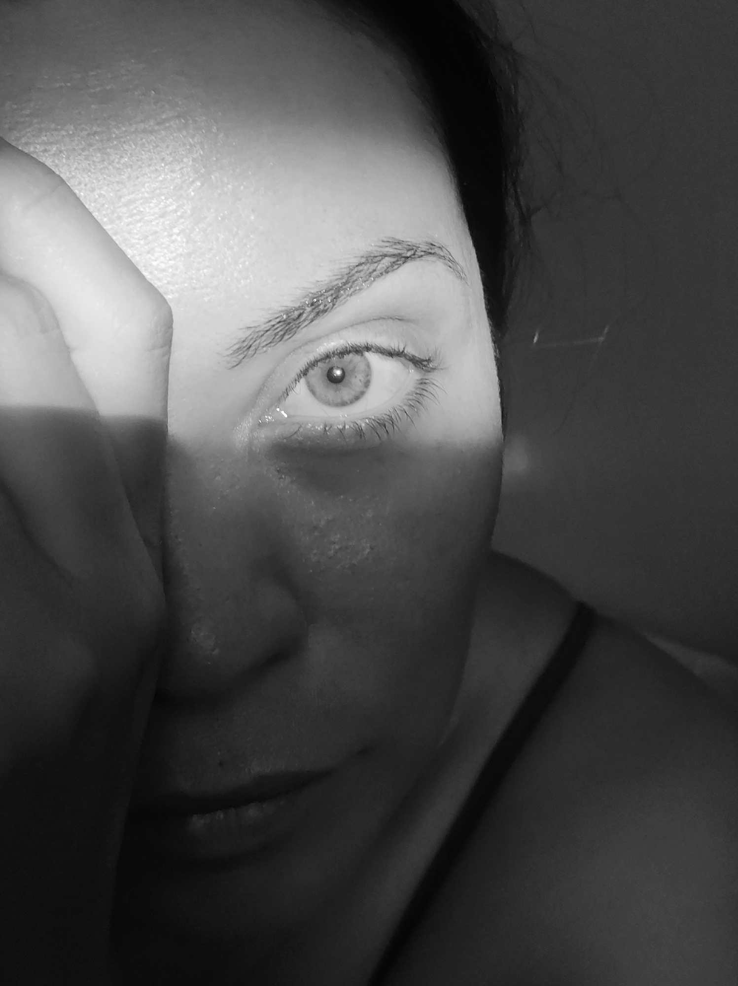 A black and white close-up photograph shows the right side of a person's face, with their right eye being the focal point. A bright stripe of light cuts vertically across their face, from forehead to cheek, obscuring parts of their nose and mouth. The person's right hand is raised to their forehead, partially covering their face and creating a shadow. Their right eye is open, with a light-colored iris and a visible pupil. Eyelashes are distinct, and their eyebrow is neatly shaped. The skin appears to have some texture, with subtle pores visible in the illuminated area. The background is dark and out of focus, suggesting an indoor setting. The lighting creates a dramatic, almost stark effect, highlighting the texture of the skin and the eye. The overall mood is somewhat enigmatic, due to the partial obscuring of the face and the dramatic lighting. No text is visible in the image.
