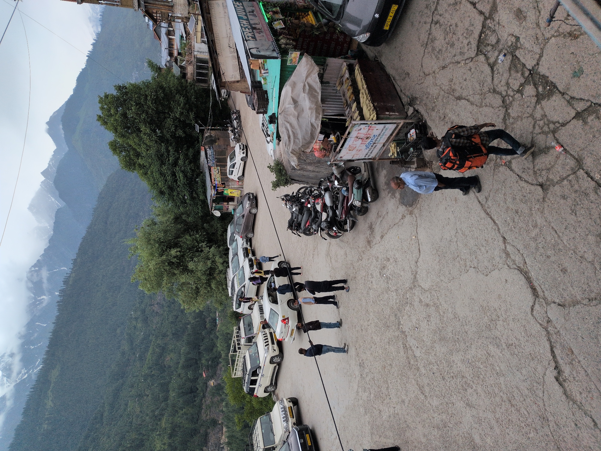 The image captures a street scene in Vashisht, India, with a backdrop of lush green mountains under a cloudy sky. In the foreground, a paved area serves as parking for several vehicles, including white SUVs, sedans, and a pickup truck. A group of individuals, seemingly tourists, are engaged in conversation near the parked cars. To the right, a vibrant stall sells fresh juice and produce, attracting attention with its colorful displays. Motorcycles are neatly parked nearby, suggesting a popular mode of transportation in the area. The overall atmosphere is one of a bustling mountain town, with a blend of local life and tourist activity.